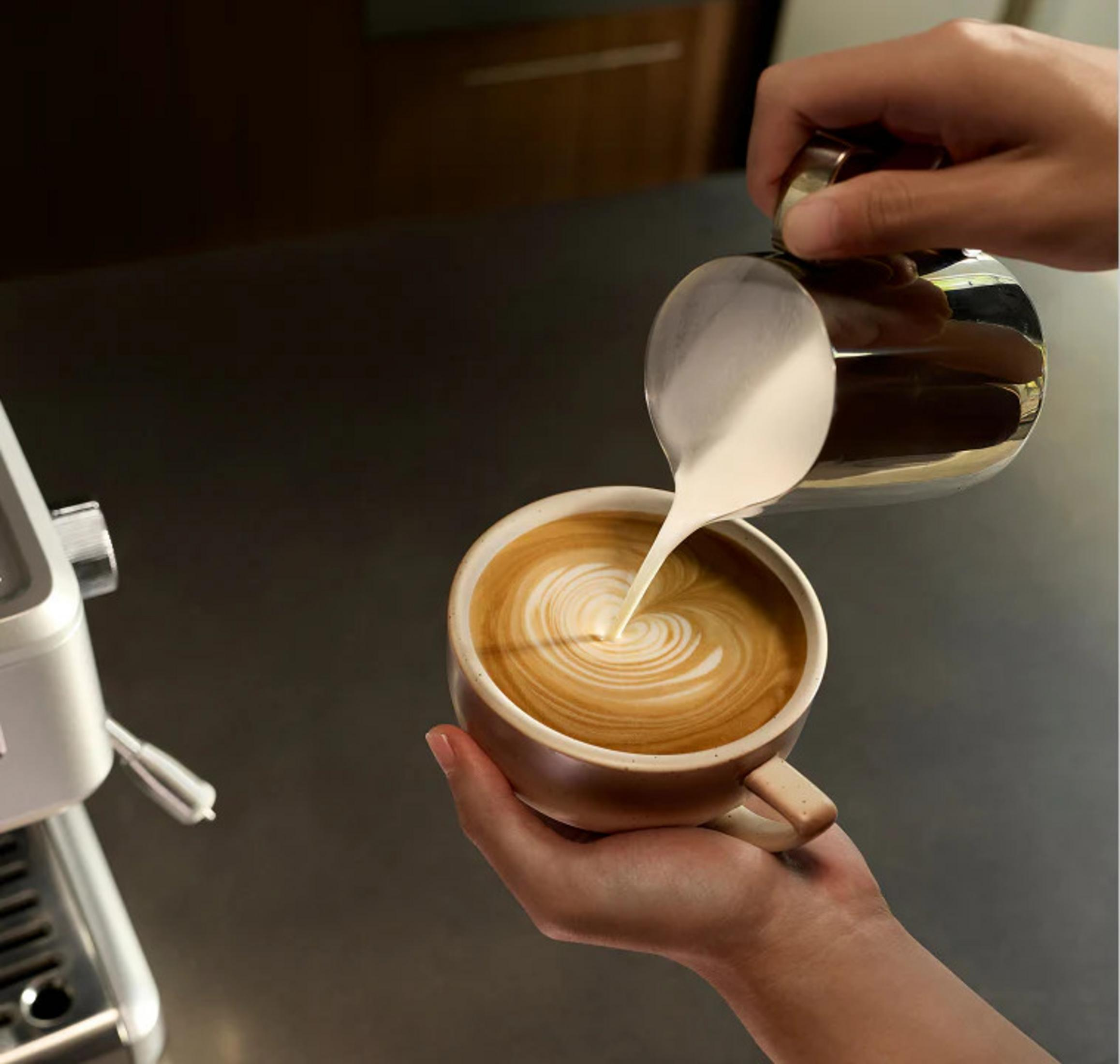 Qualcuno versa il latte da una lattiera di metallo in una tazza di caffè, creando latte art.
