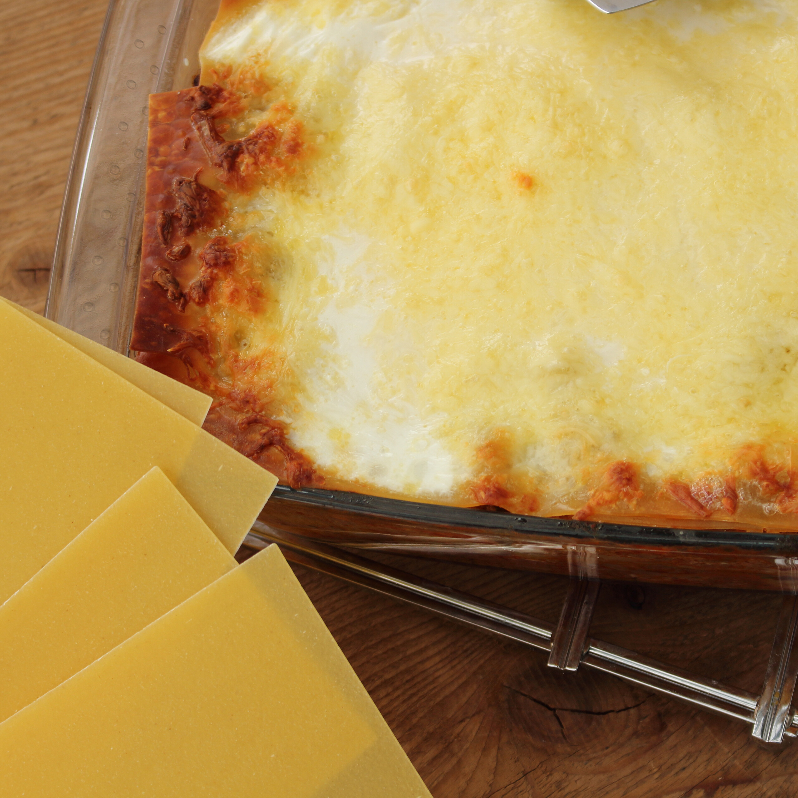 Frisch gebackene Lasagne in einer Glasauflaufform mit rohen Lasagnenudeln daneben auf einem Holzbrett.