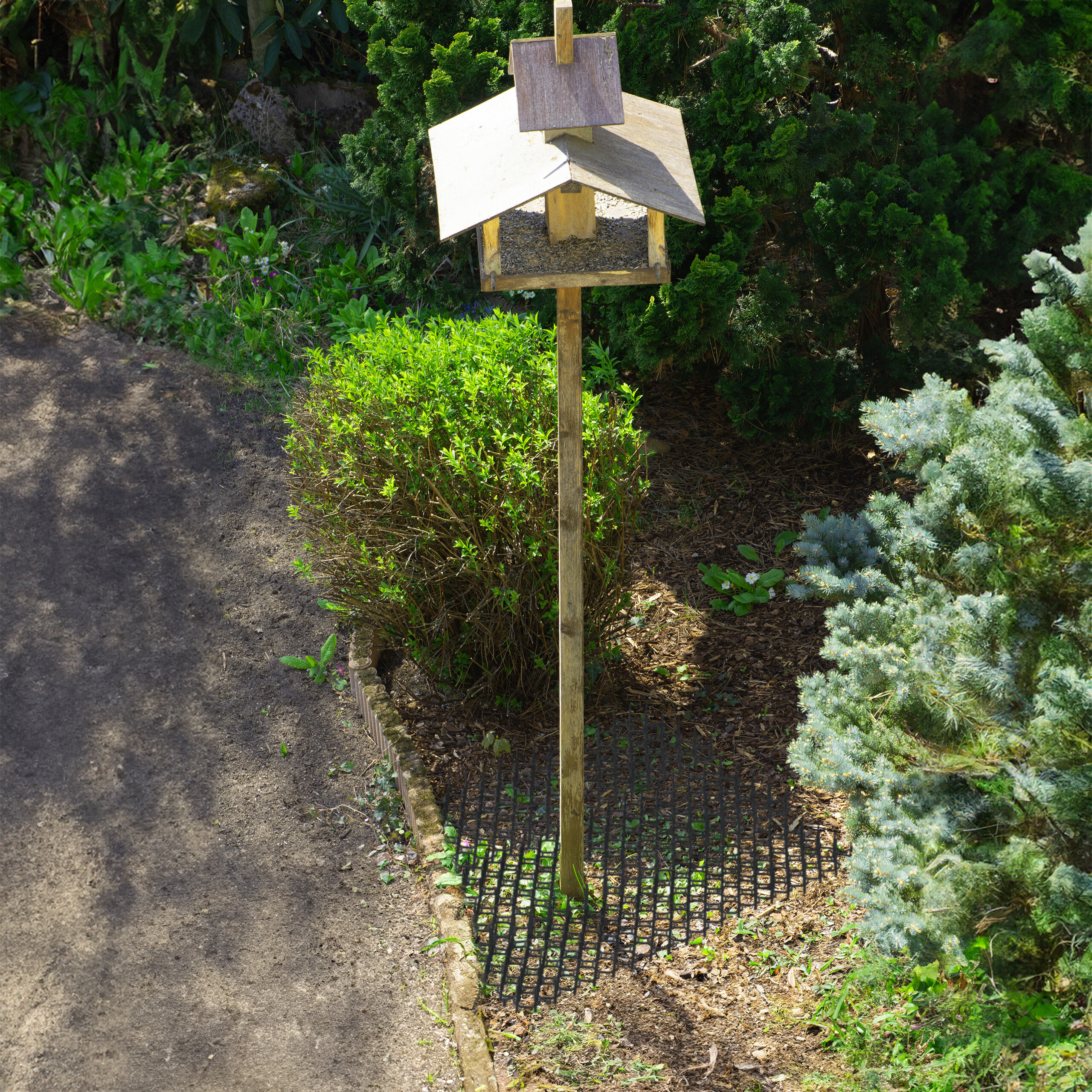 Ein hölzernes Vogelhäuschen auf einem langen Pfahl im Garten neben einem Weg und Büschen.
