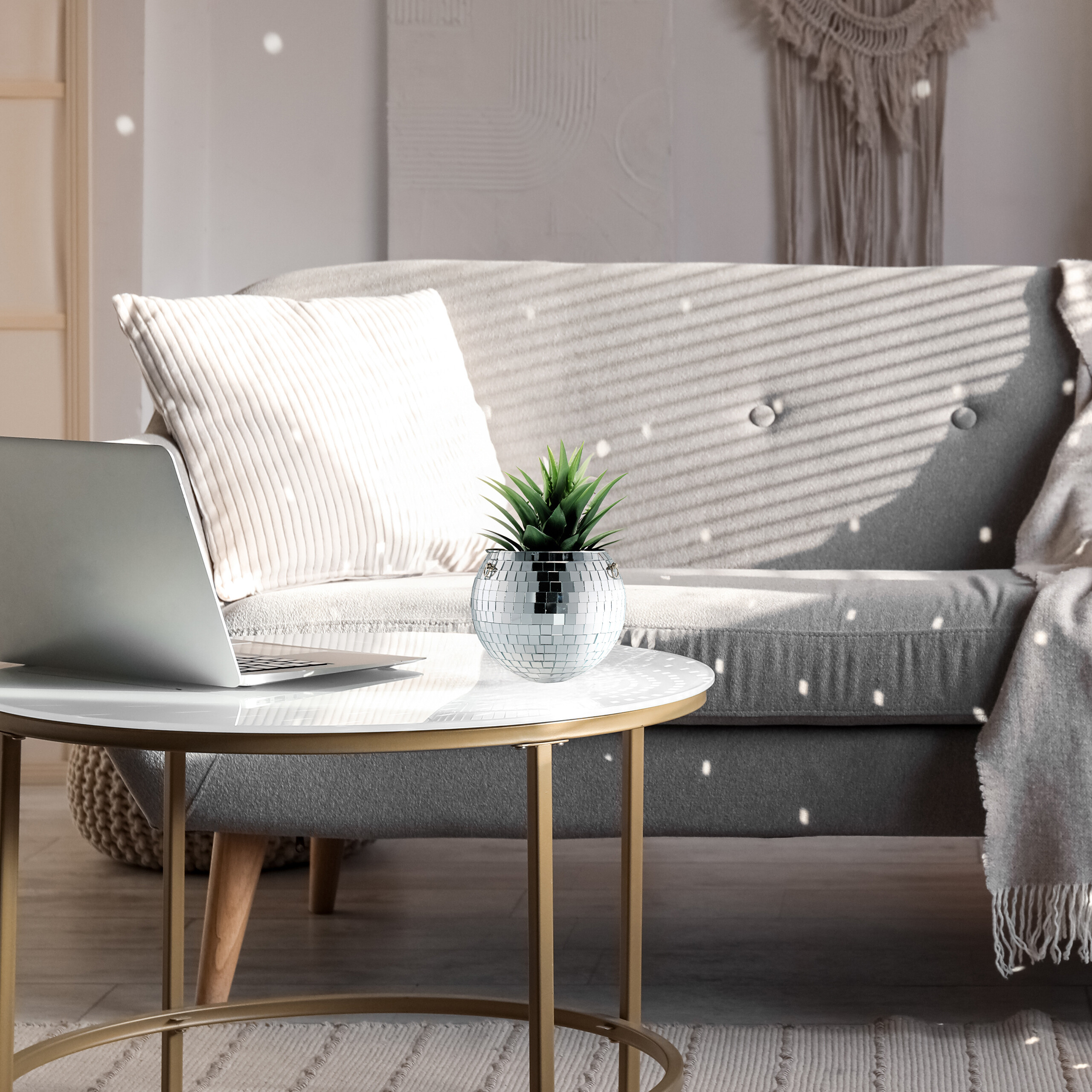 Ein Laptop und ein glitzernder Übertopf mit Pflanze stehen auf einem Couchtisch vor einem grauen Sofa.