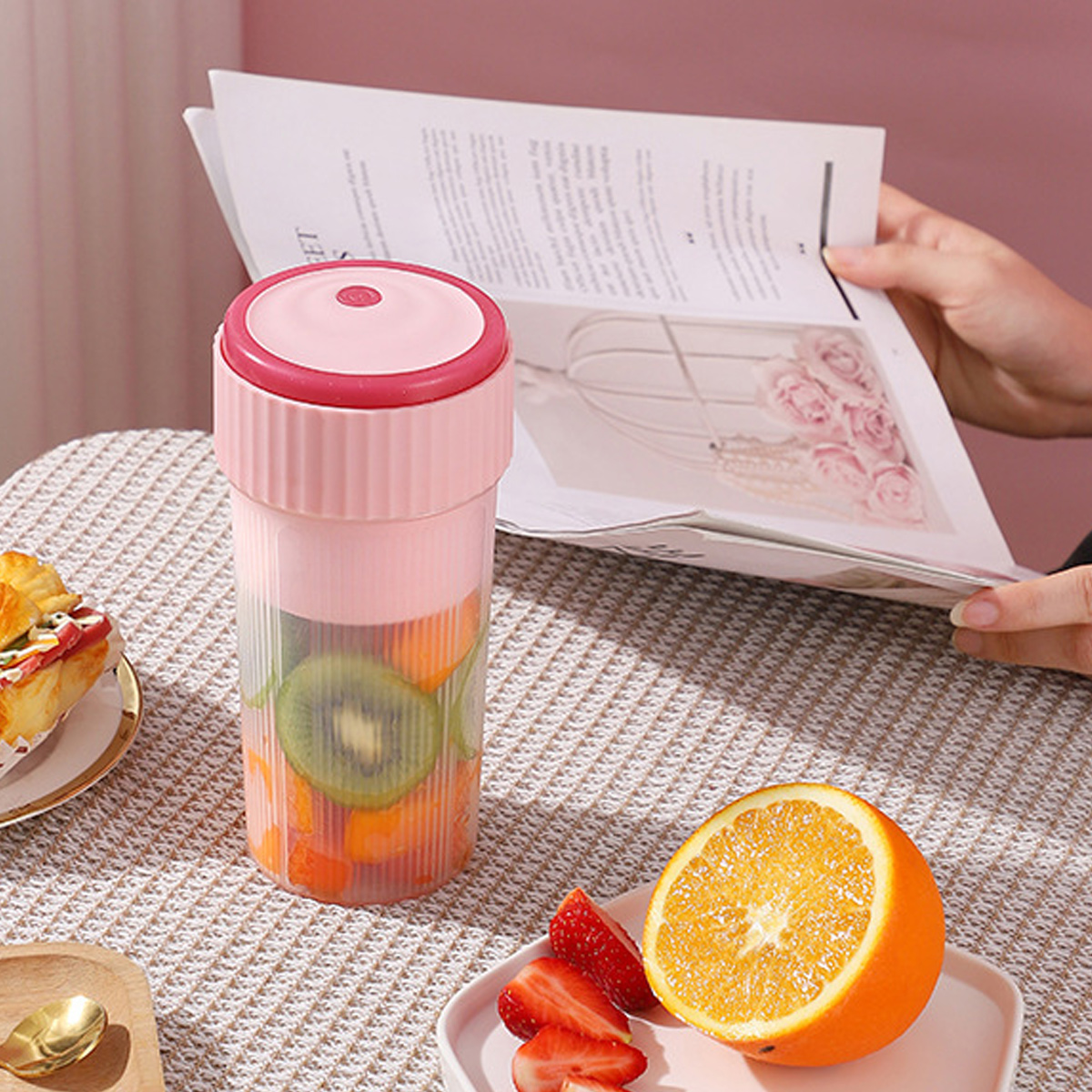 Ein rosa tragbarer Mixer mit Obst, eine Orangenhälfte und Erdbeeren auf einem Tisch mit einer Zeitschrift im Hintergrund.