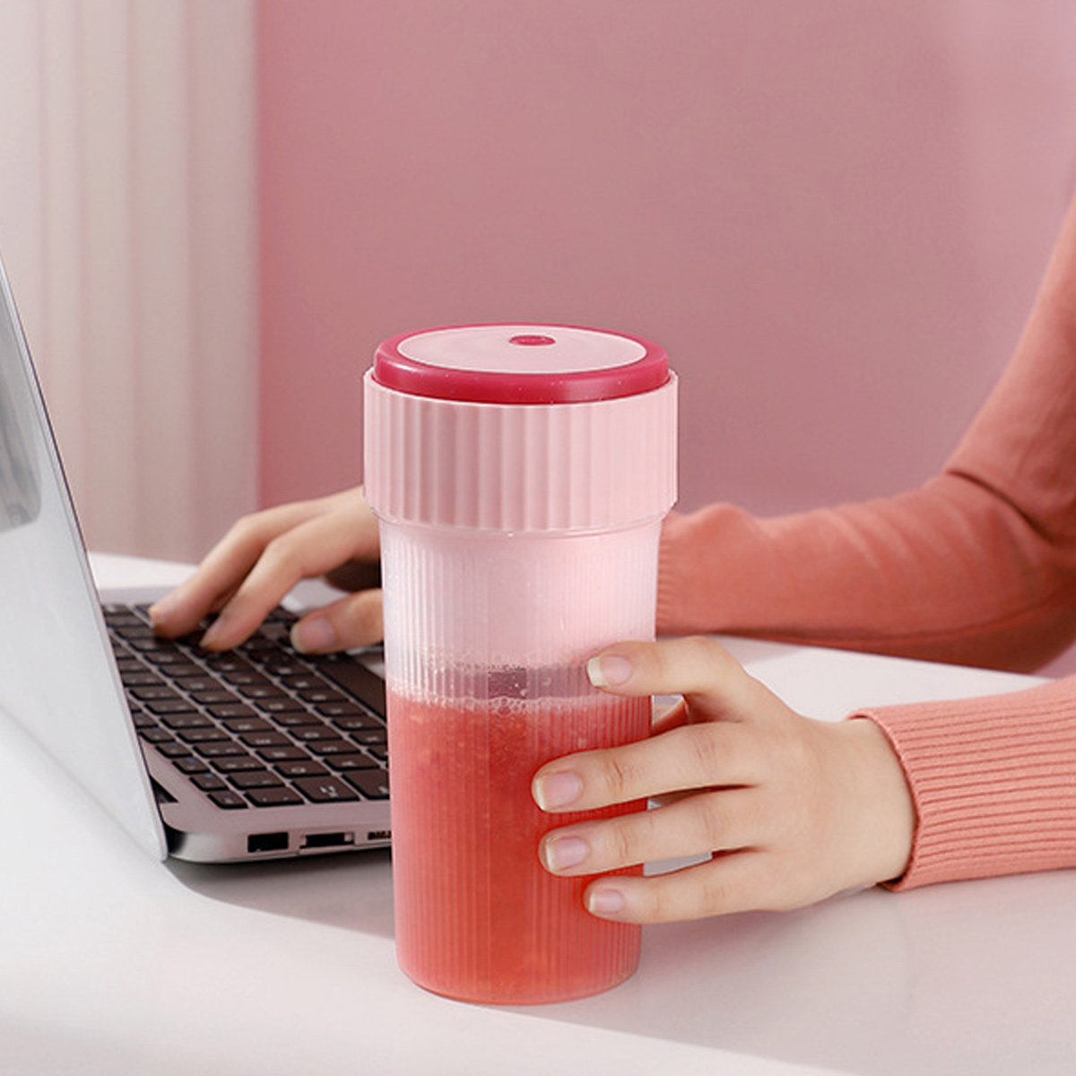 Person in rosafarbenem Oberteil hält einen rosa Smoothie-Becher neben einem Laptop auf einem Tisch.