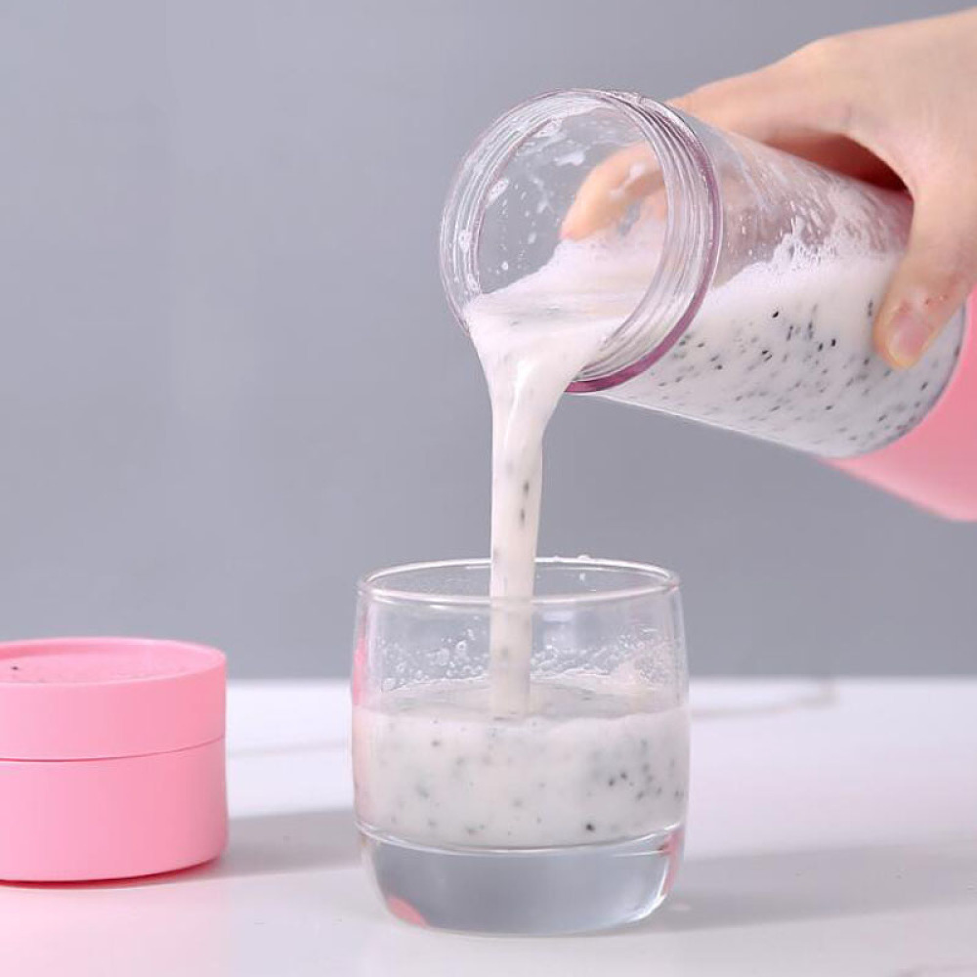 Eine Hand gießt einen weißen Smoothie mit schwarzen Samen aus einer rosa Mixerflasche in ein Glas.
