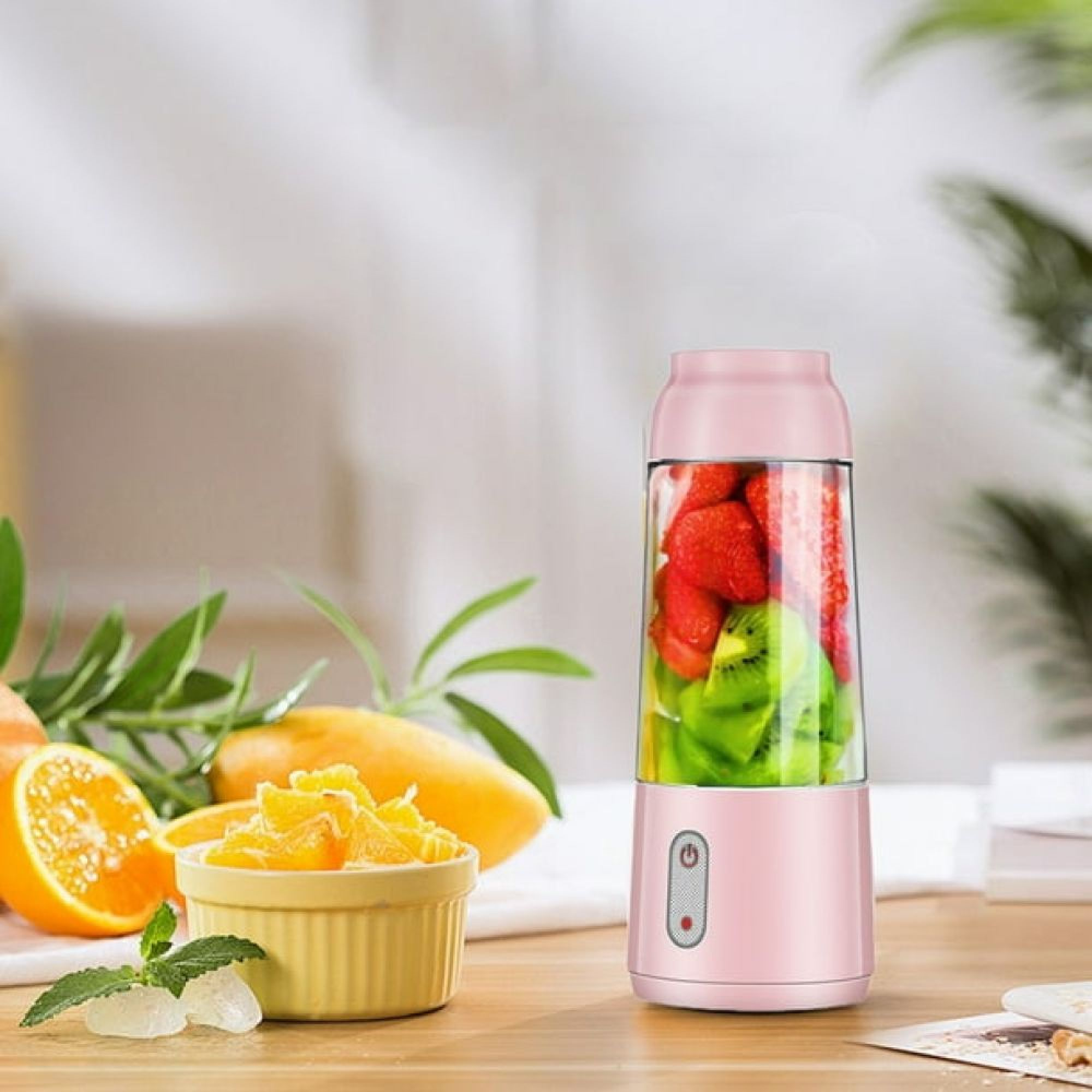 Ein rosa tragbarer Mixer mit Erdbeeren und Kiwis steht auf einem Holztisch neben Orangenscheiben und einer Mango.