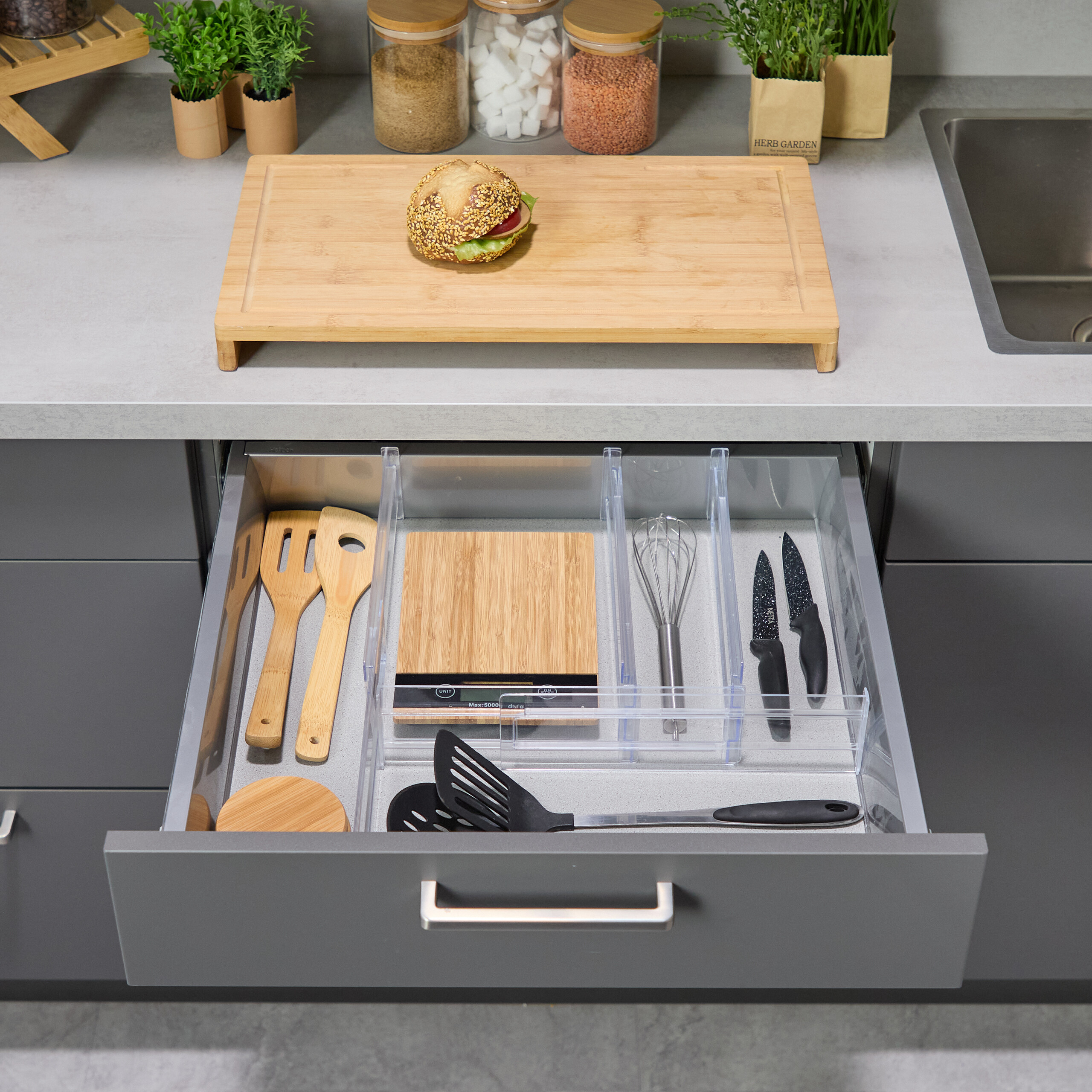 Eine offene Küchenschublade mit Utensilien, Küchenwaage, Schneebesen und Messern unter einer Theke mit Sandwich.