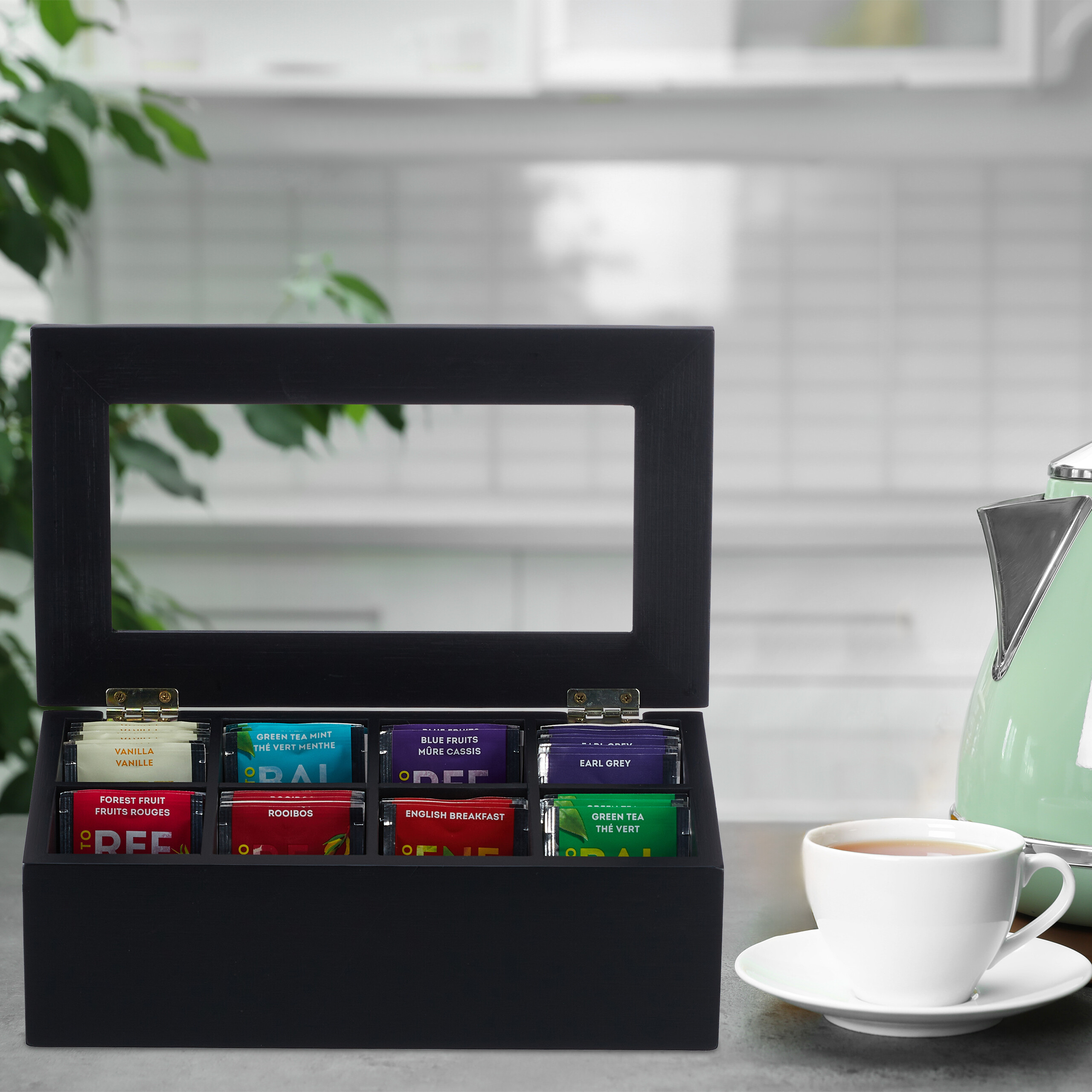 Eine offene schwarze Teebox mit bunten Teebeuteln, neben einer weiße Tasse und einem hellgrünen Wasserkocher.