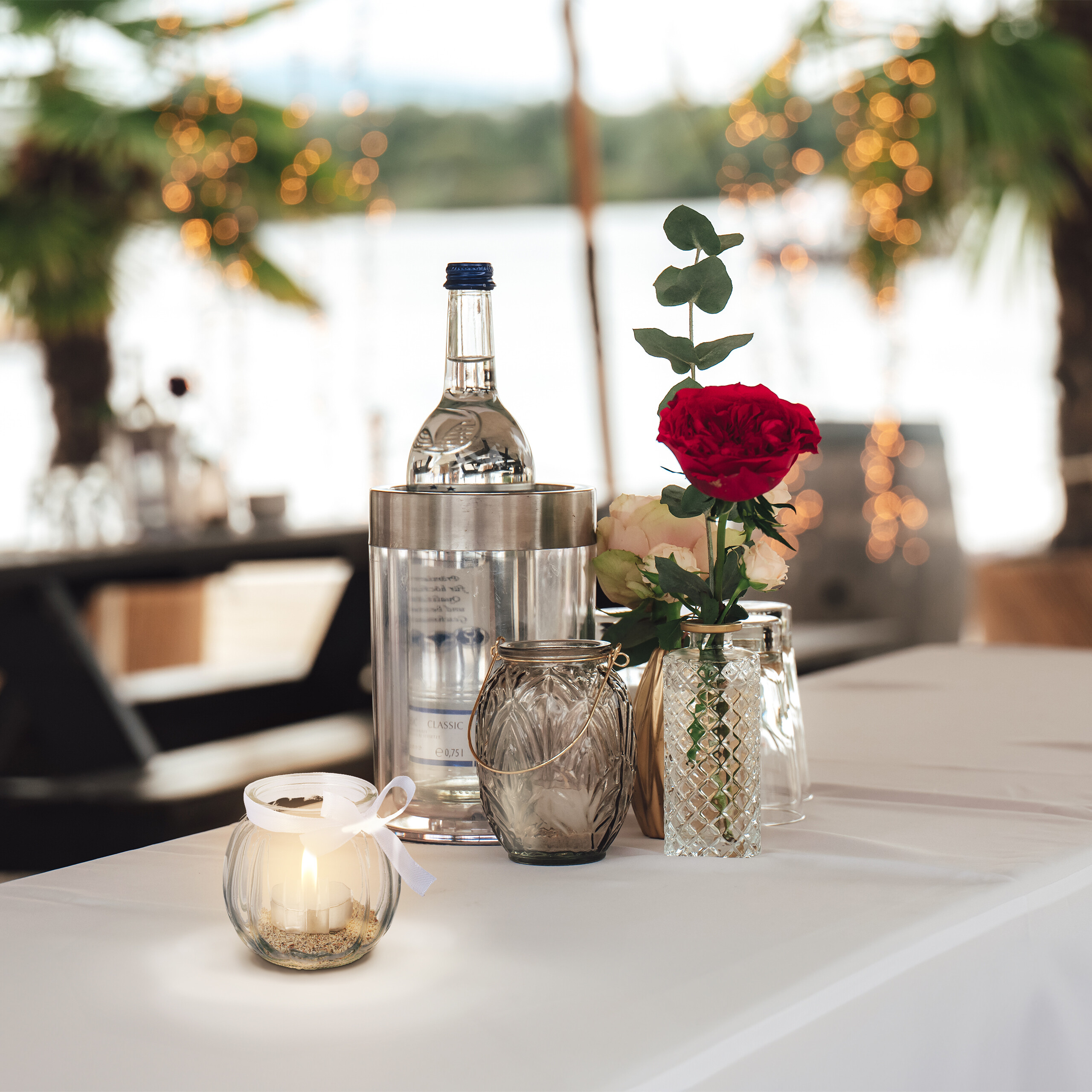 Ein Tisch mit einer Flasche in einem Eisk übel, einer roten Rose in einer Vase und brennenden Kerzen im Freien.