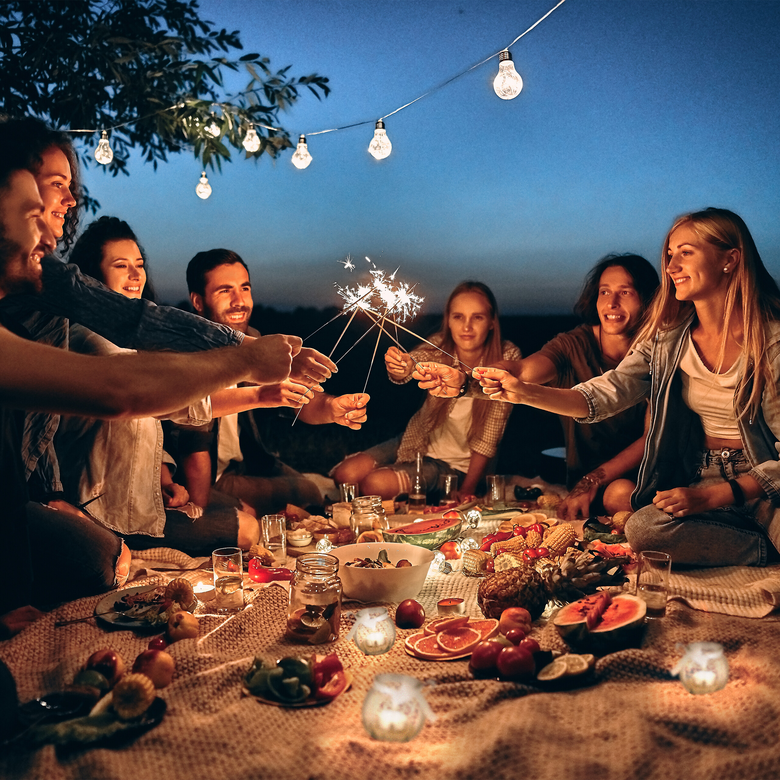 Freunde halten nachts bei einem Picknick mit Lichtern Wunderkerzen in die Höhe.