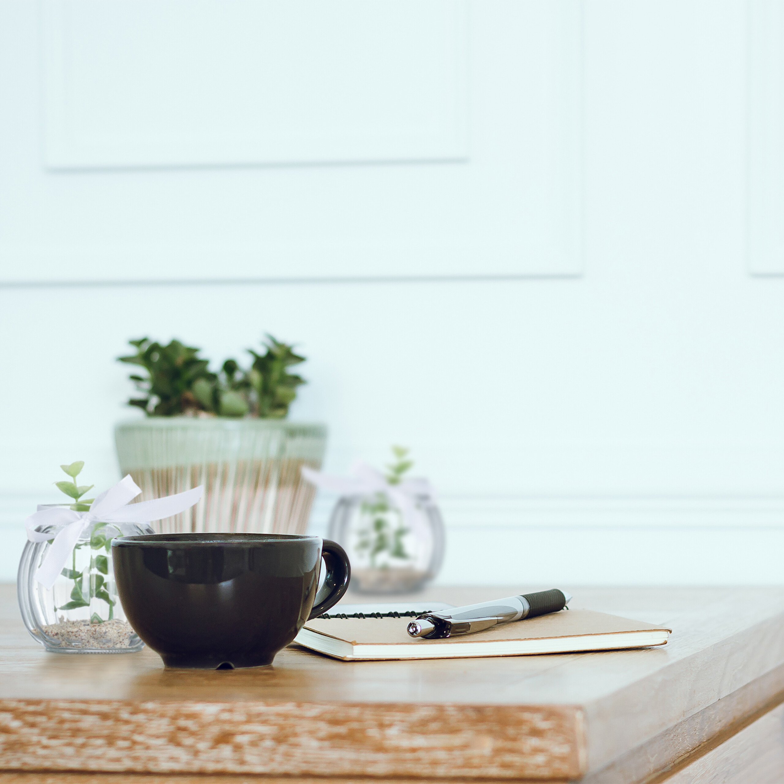Eine schwarze Tasse, ein Notizbuch und ein Stift auf einem Holztisch vor einer hellen Wand mit kleinen Pflanzen.