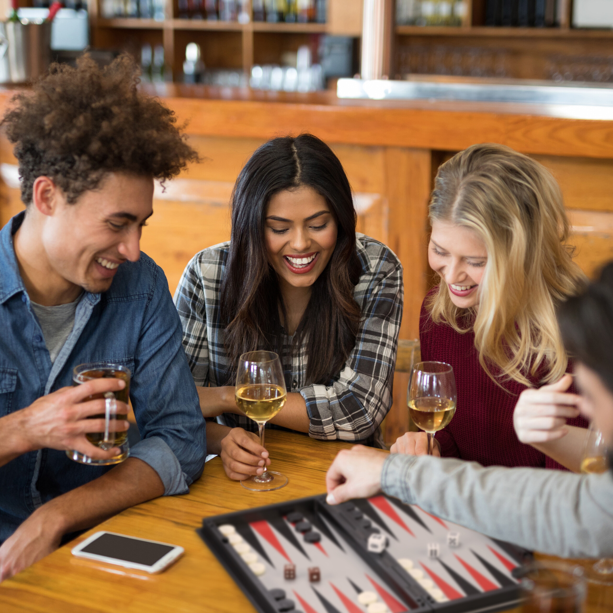 Freunde lachen und spielen Backgammon an einem Holztisch in einer Bar und halten Gläser mit Bier und Wein.