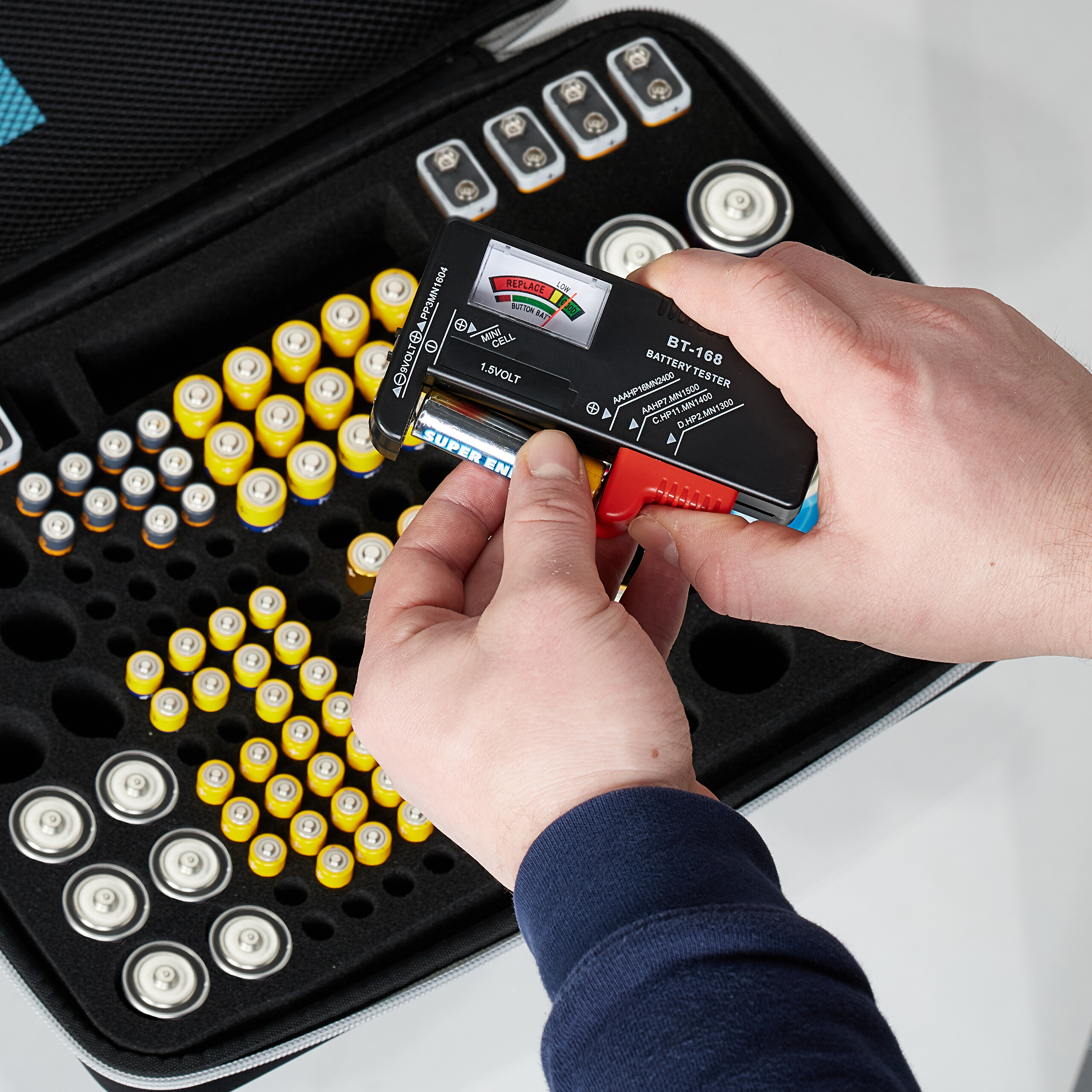 Ein Hände testen eine Batterie in einem BT-168 Tester über einer aufrechten Schaumstoffbox voller Batterien.