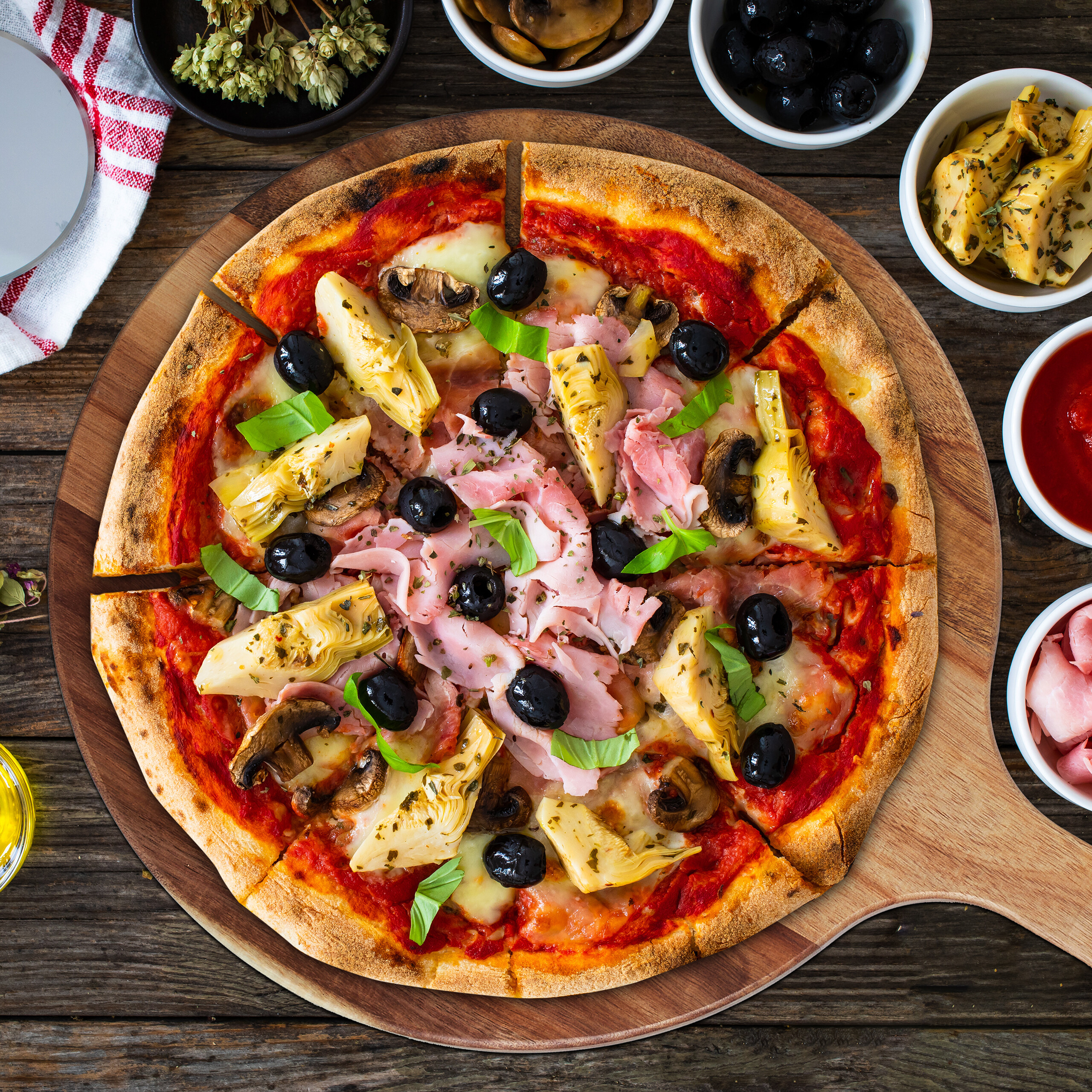 Eine Pizza mit Schinken, Artischocken, Pilzen, Oliven und Basilikum auf einem Holzbrett, umgeben von Schüsseln.
