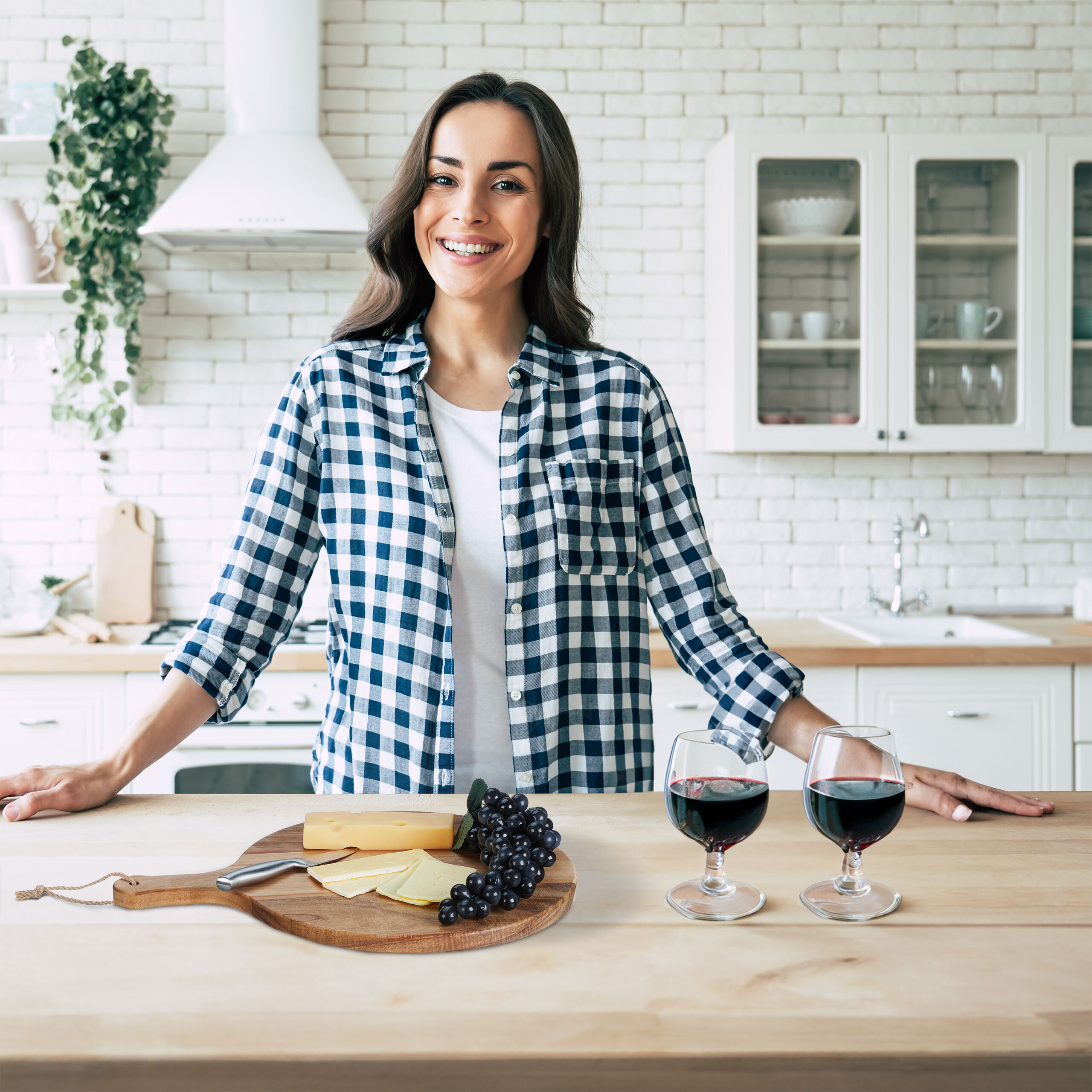 Eine lächelnde Frau steht in einer Küche an einer Holztheke mit Käse, Trauben und zwei Gläsern Rotwein.