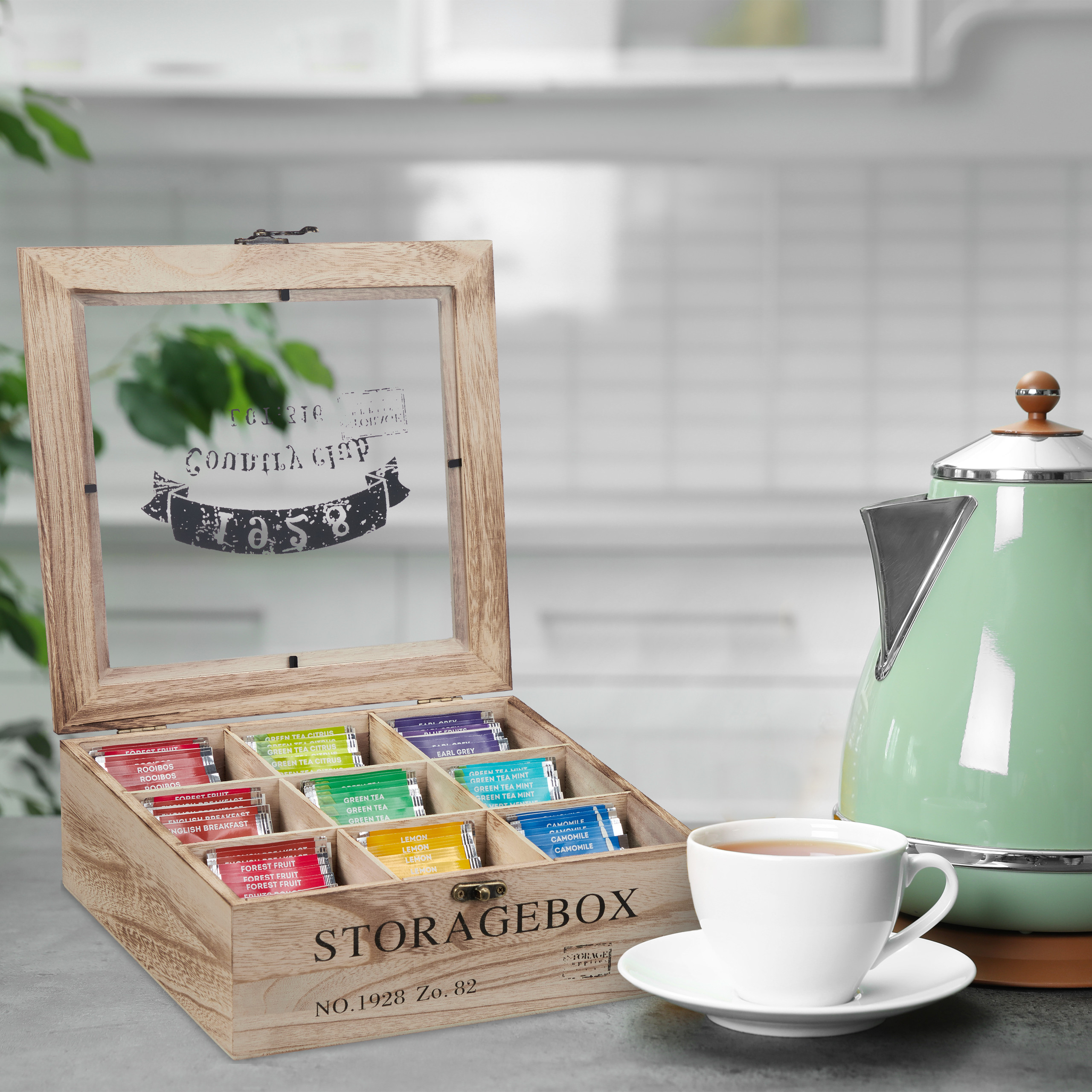 Eine offene Teebox aus Holz mit bunten Teebeuteln, eine weiße Tasse auf Untertasse und ein grüner Wasserkocher.