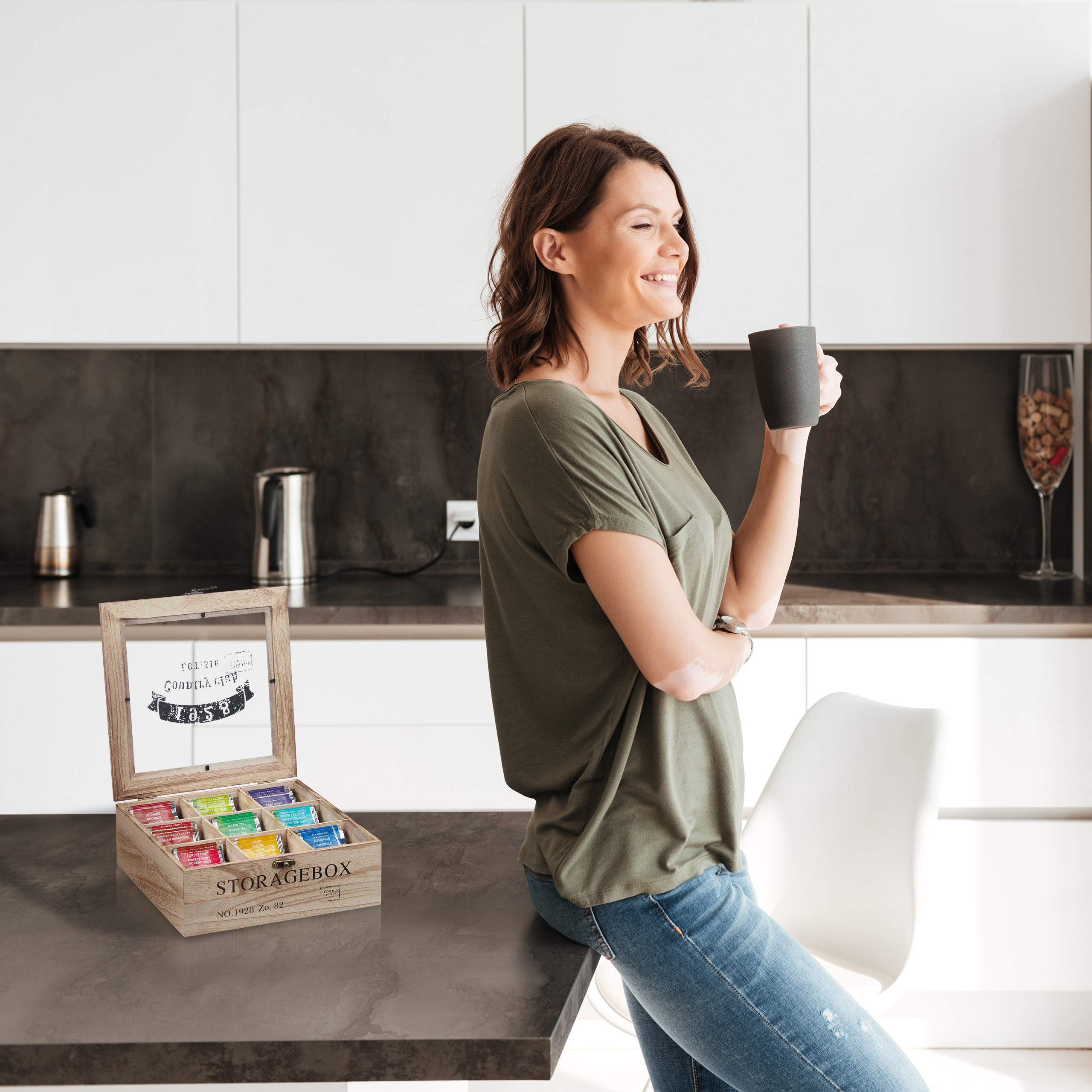 Eine lächelnde Frau lehnt an einer Küchentheke, hält eine Tasse und blickt auf eine hölzerne Tee-Aufbewahrungsbox.