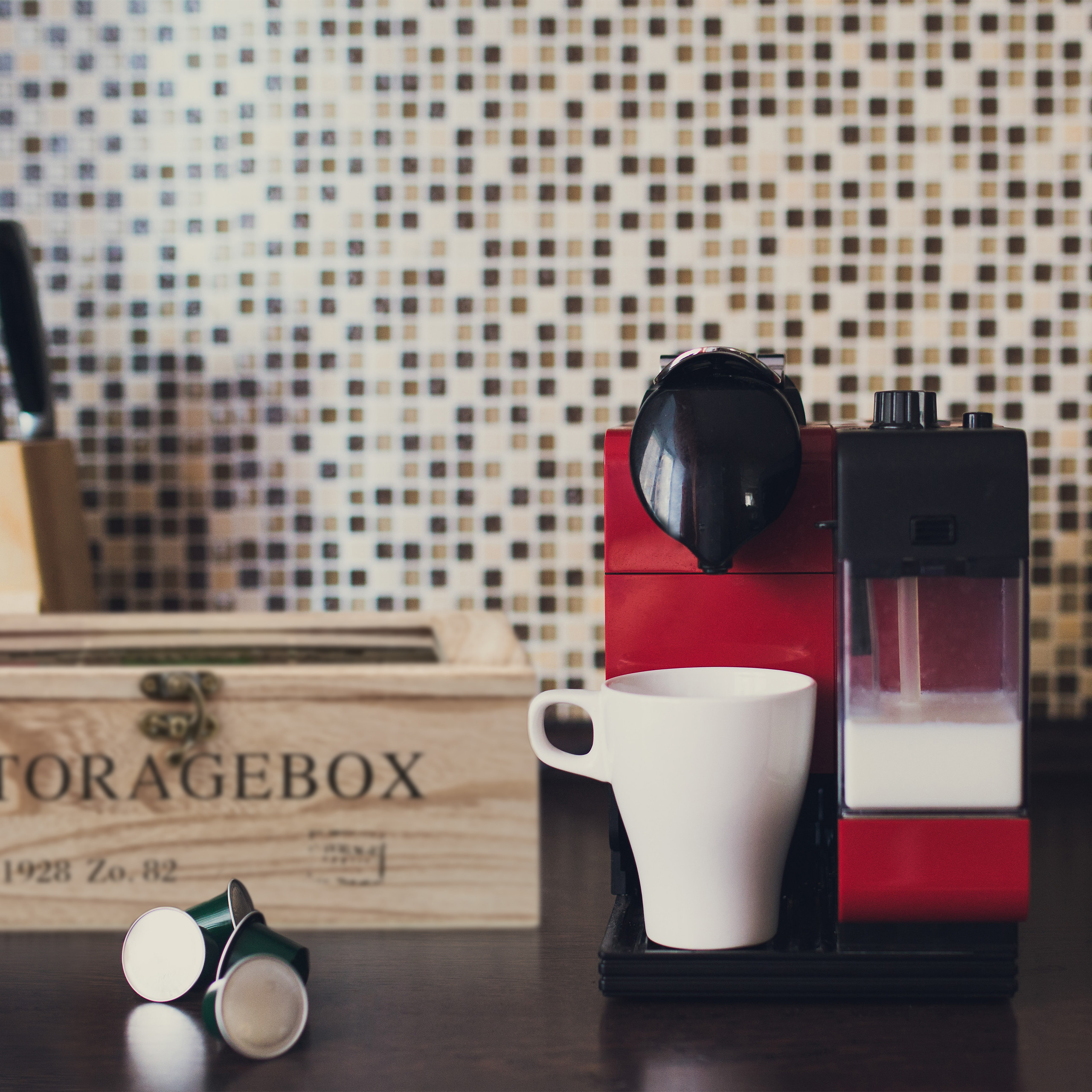 Eine rote und schwarze Kaffeemaschine mit weißer Tasse, Kapseln und Kiste auf dunkler Theke vor Fliesenhintergrund.