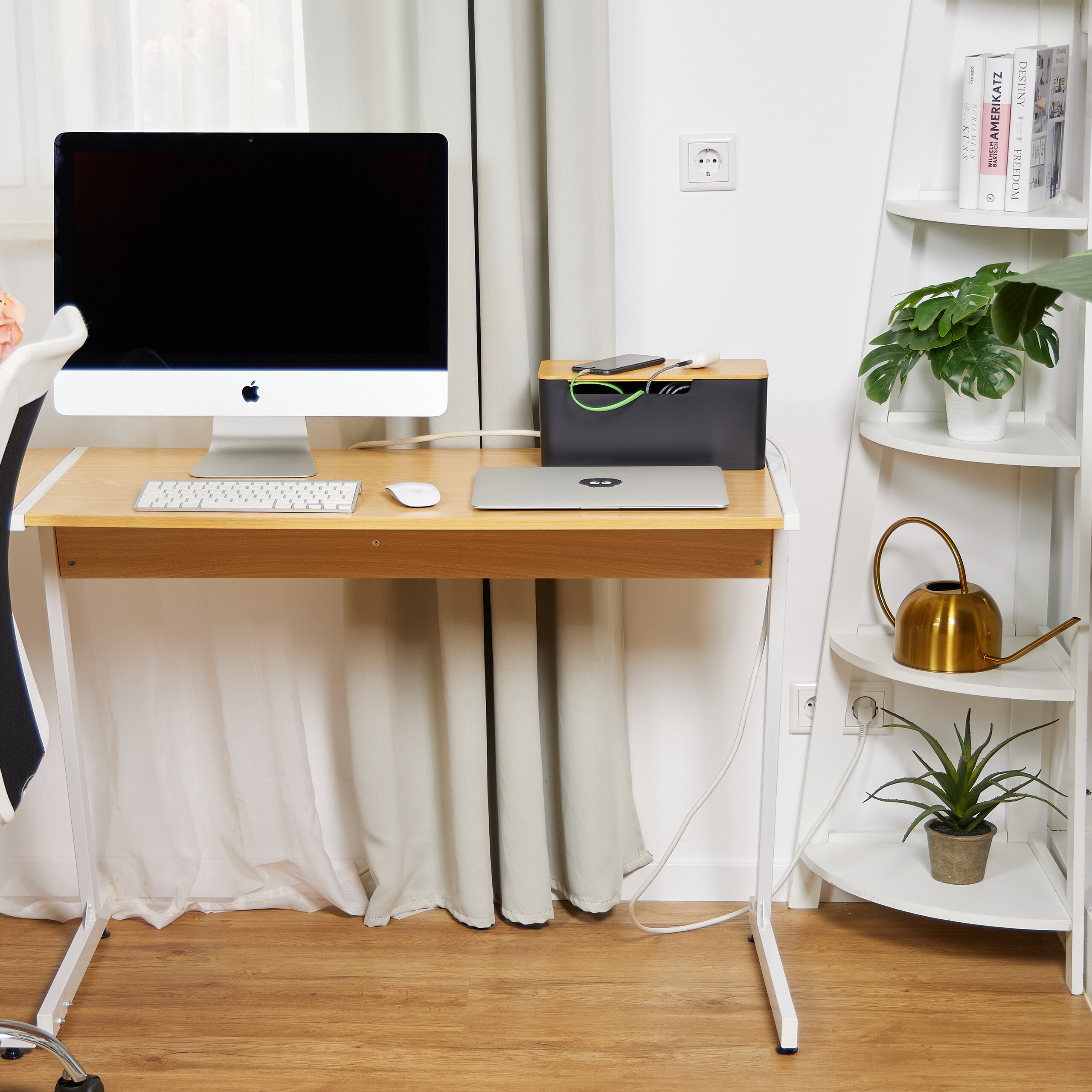 Ein Holztreibtisch mit Computer, Laptop und Zubehoer. Rechts steht ein weisses Regal mit Pflanzen.