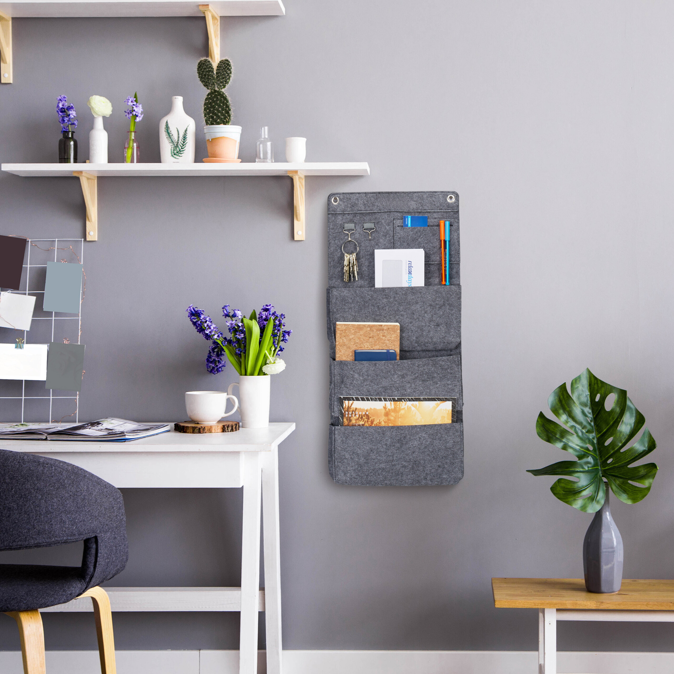 Grauer Filz-Wandorganizer an grauer Wand über Schreibtisch mit Blumen und Stuhl sowie Monstera-Pflanze rechts.