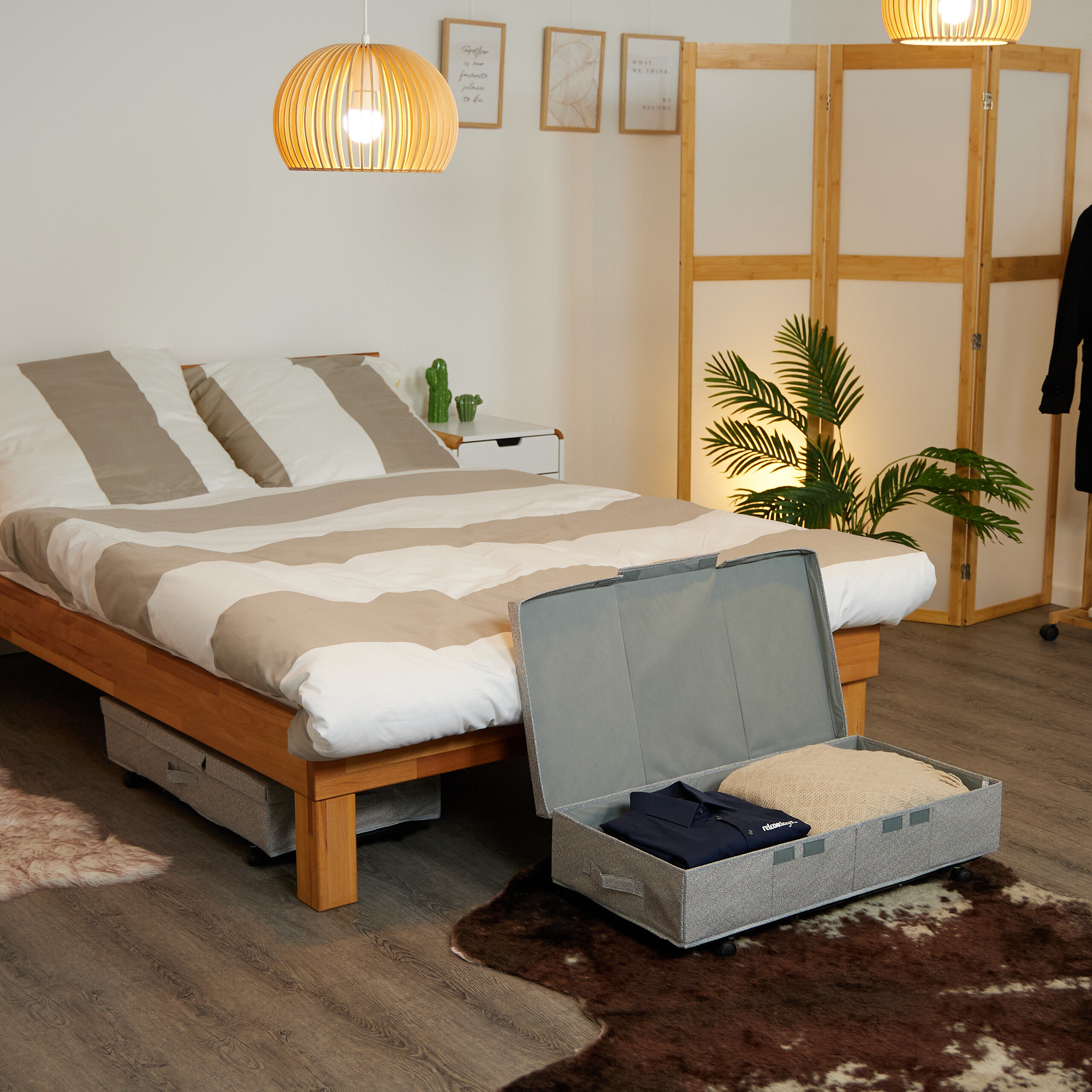 Schlafzimmer mit Holzbett, offenen grauen Kisten auf Rollen mit Kleidung, Pflanzen und dekorativen Holzlampen.
