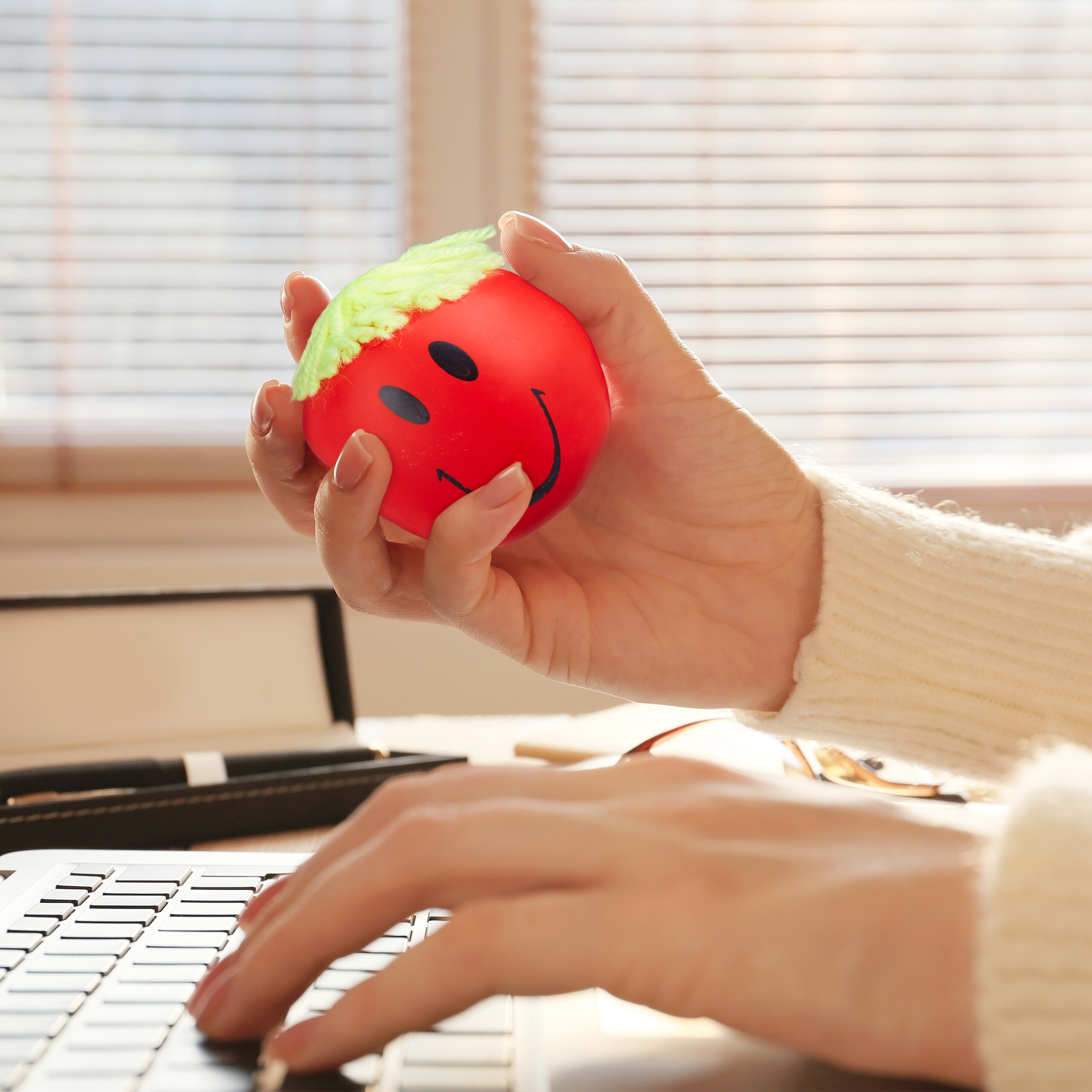 Eine Hand tippt auf einer Laptop-Tastatur, während die andere einen roten Stressball mit einem Smiley-Gesicht hält.