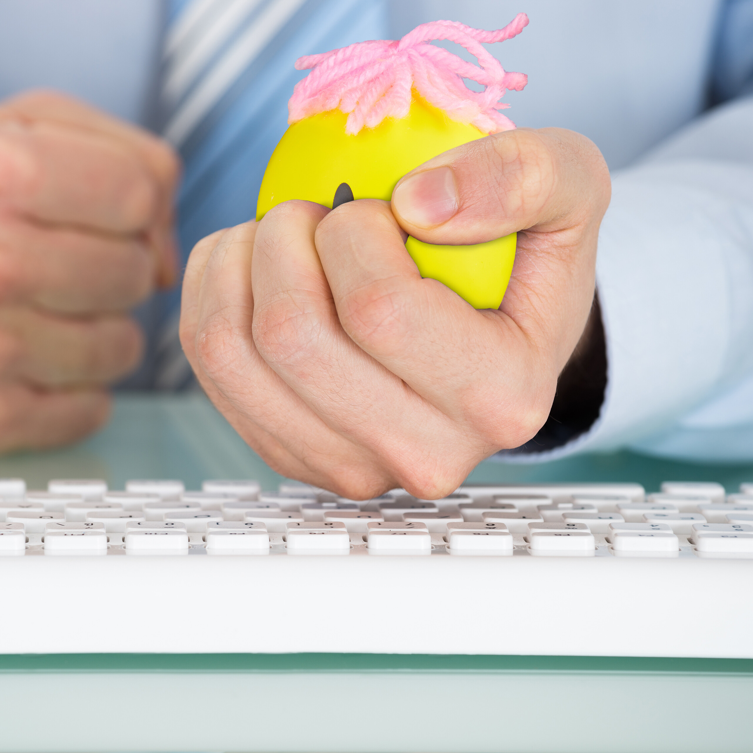 Ein Geschäftsmann drückt fest einen gelben Stressball mit rosa Haaren über einer Computertastatur.
