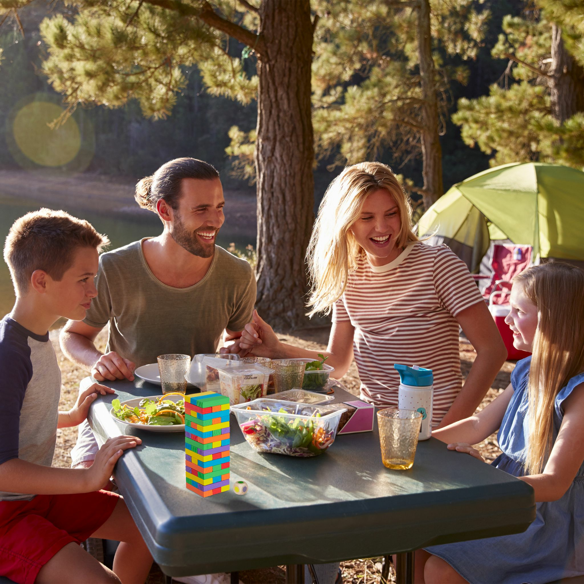 Eine Familie mit zwei Kindern sitzt an einem Picknicktisch im Freien beim Essen, im Hintergrund ist ein Zelt zu sehen.
