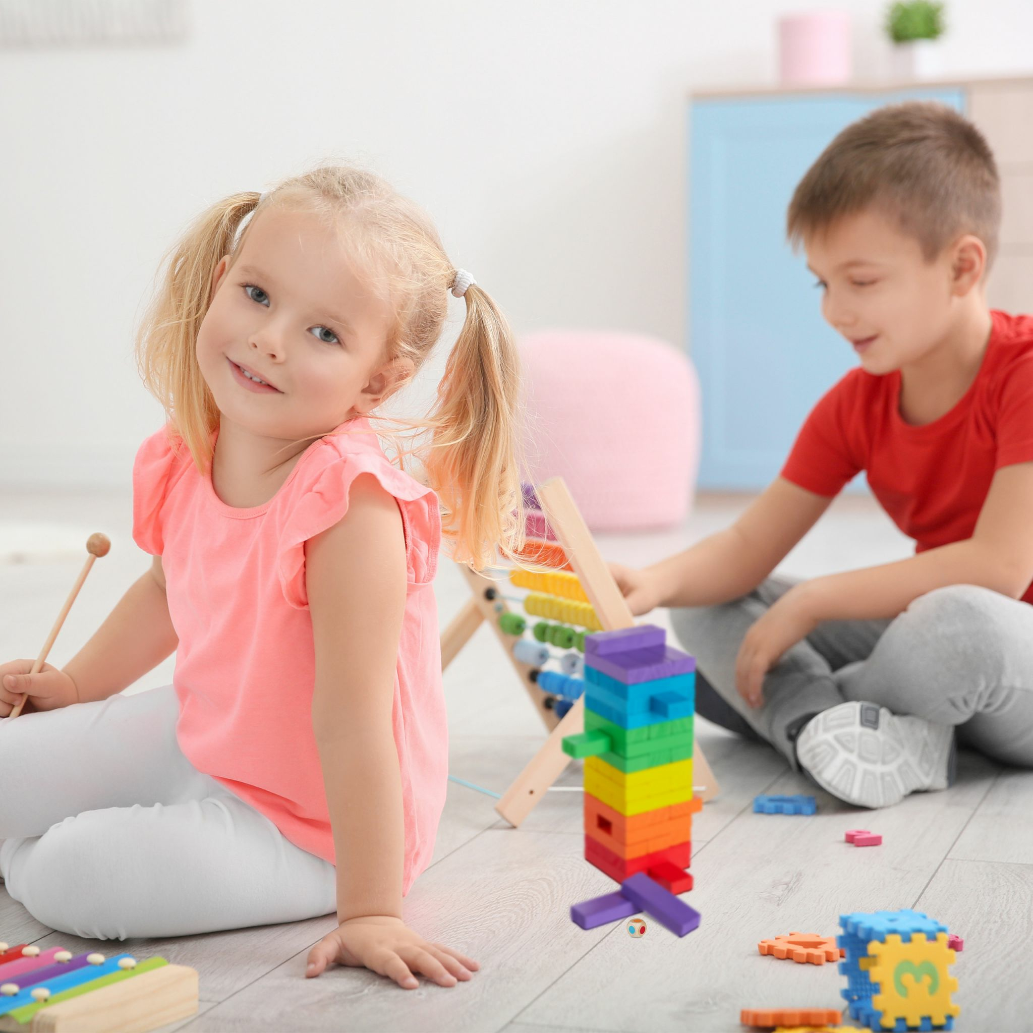 Ein blondes Mädchen in Rosa sitzt mit Spielzeug auf dem Boden, während im Hintergrund ein Junge spielt.