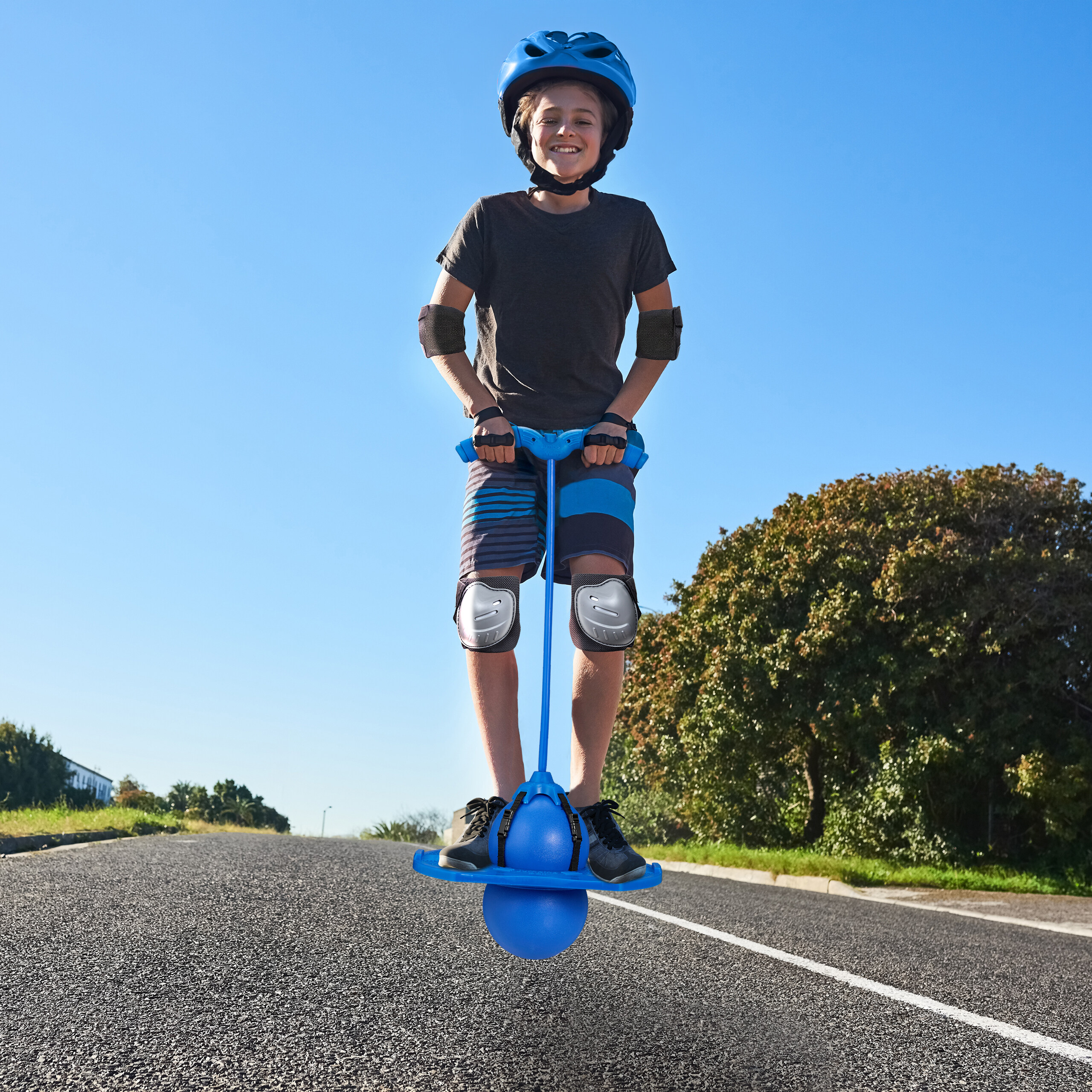 Ein Junge mit Helm und Schutzkleidung hüpft auf einem blauen Pogo-Ball auf einer Asphaltstraße.