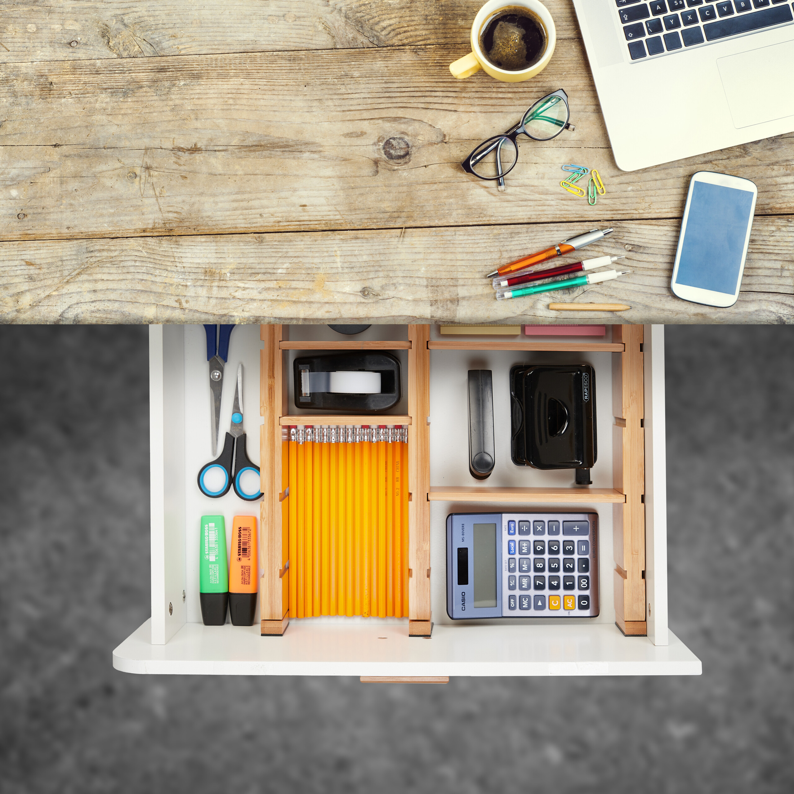Ein Holztisch mit Laptop, Tasse, Smartphone und einer geöffneten Schublade voller Büroartikel.