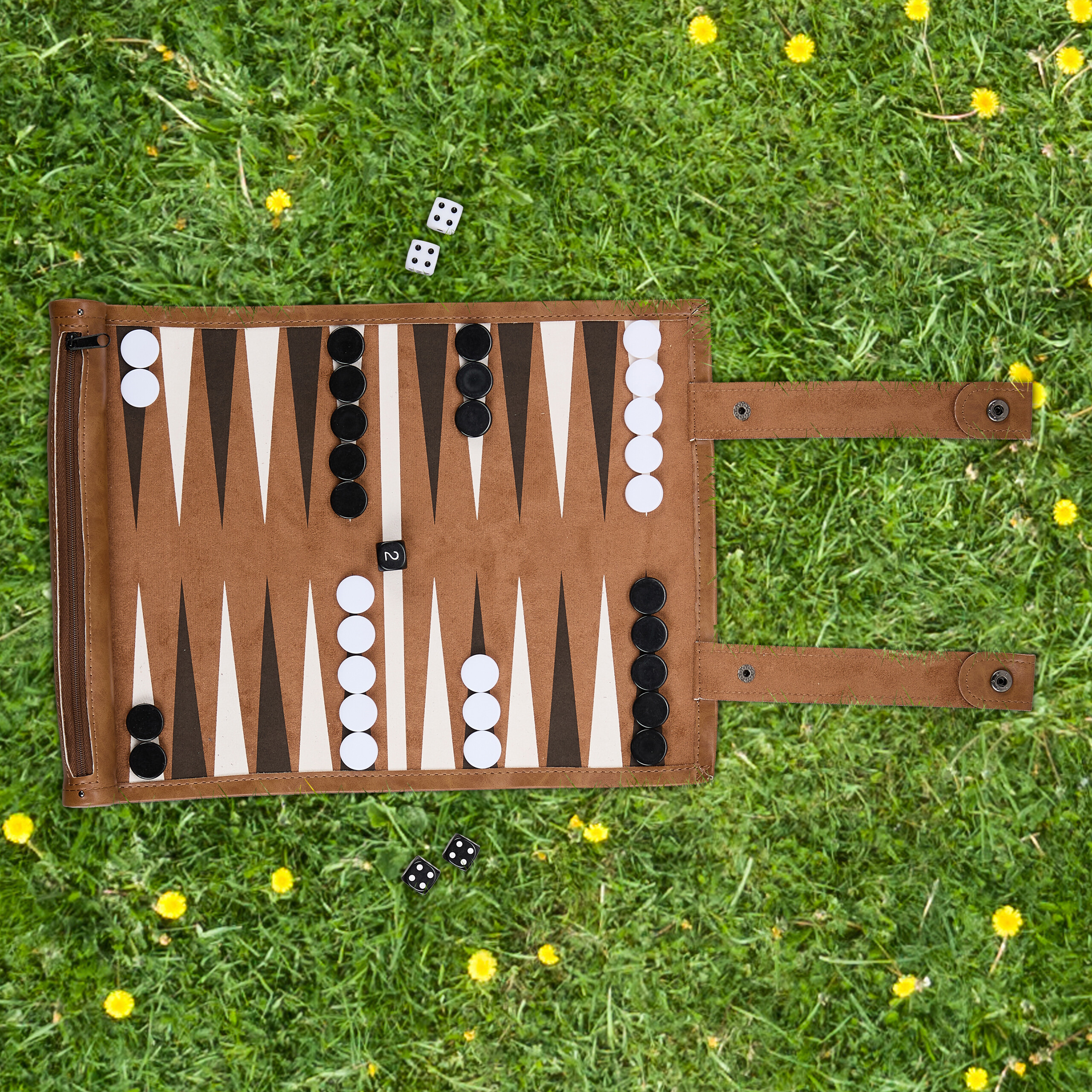 Ein Backgammon-Spielset mit braunem Lederimitat liegt auf grüner Wiese mit kleinen gelben Blumen.