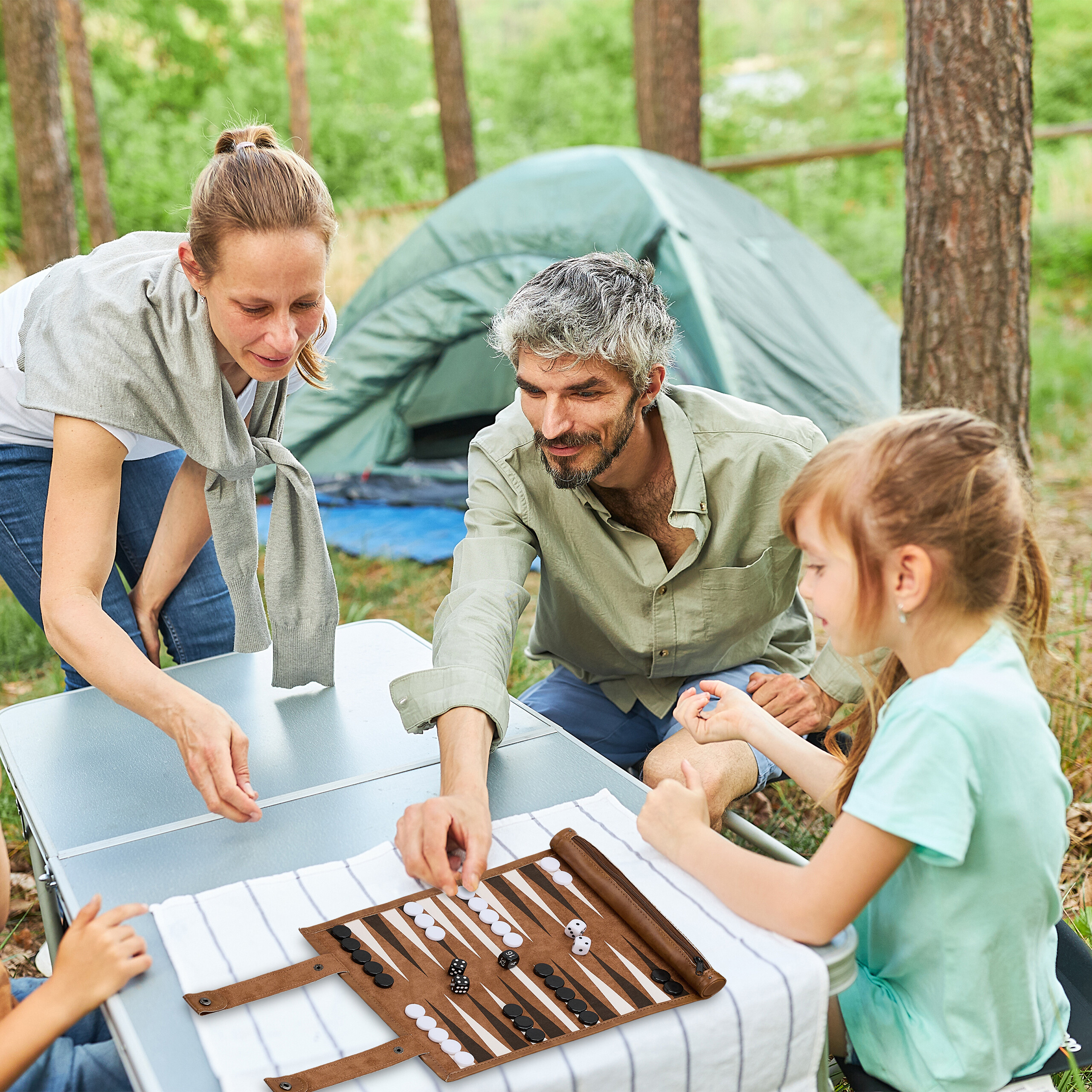 Eine dreiköpfige Familie spielt draußen in einem Wald in der Nähe eines grünen Zeltes Backgammon.