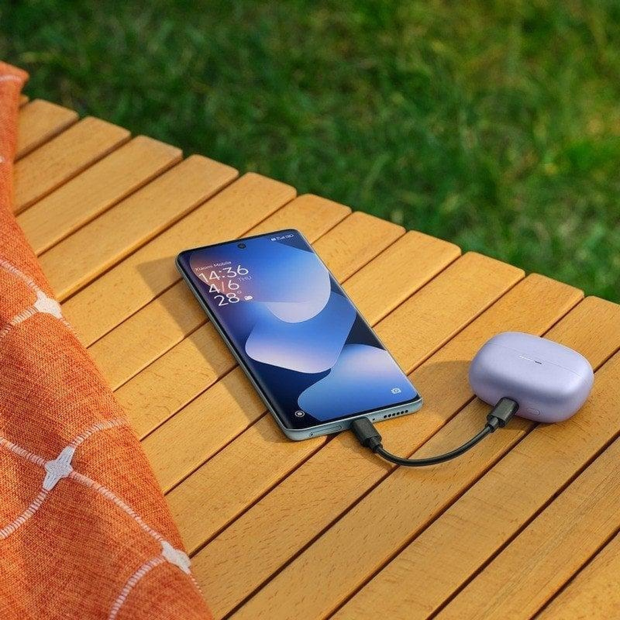 Uno smartphone e una custodia di ricarica per auricolari, collegati da un cavo, su un tavolo di legno all'aperto.