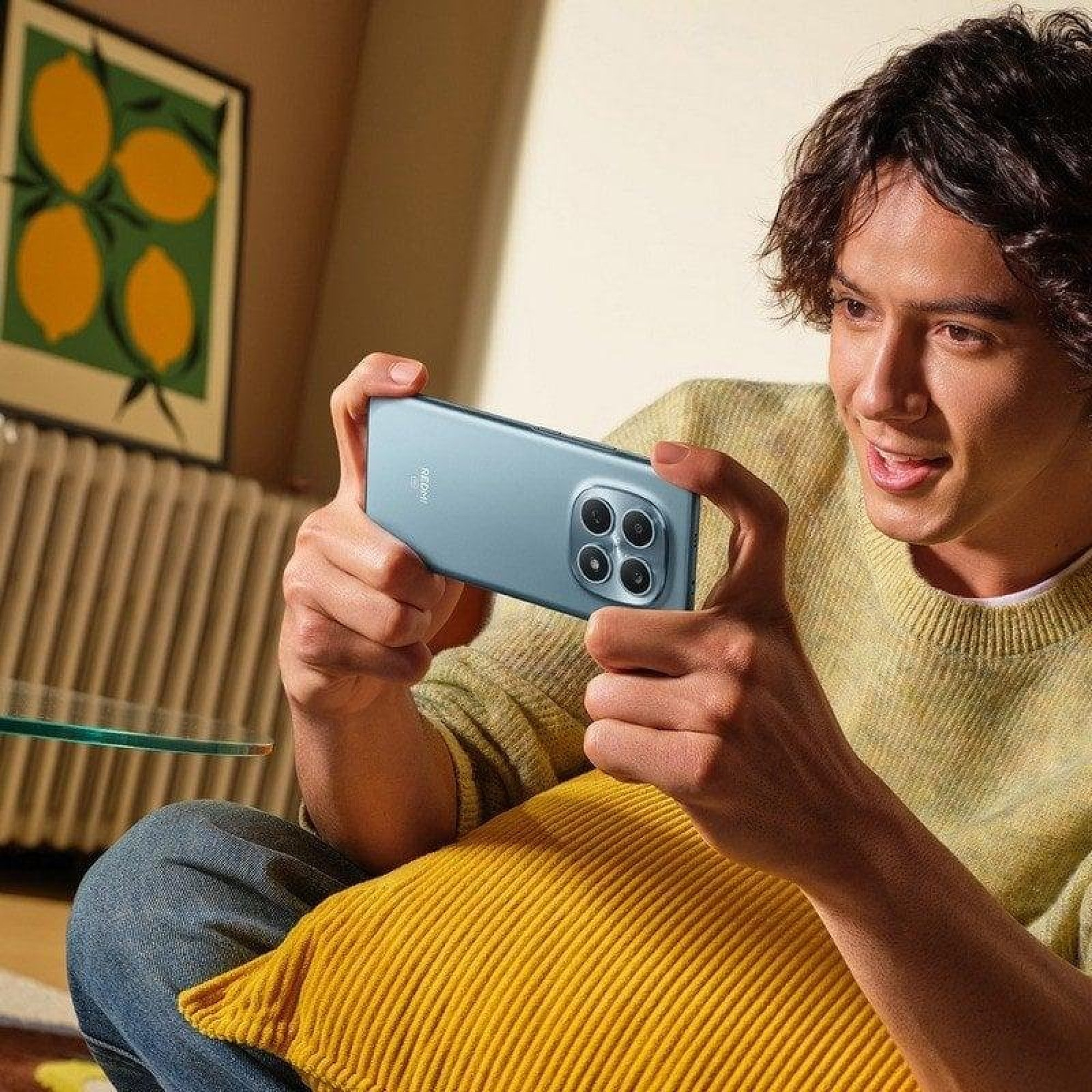 Un giovane con un maglione giallo è seduto su un cuscino e tiene in mano uno smartphone azzurro in orizzontale.