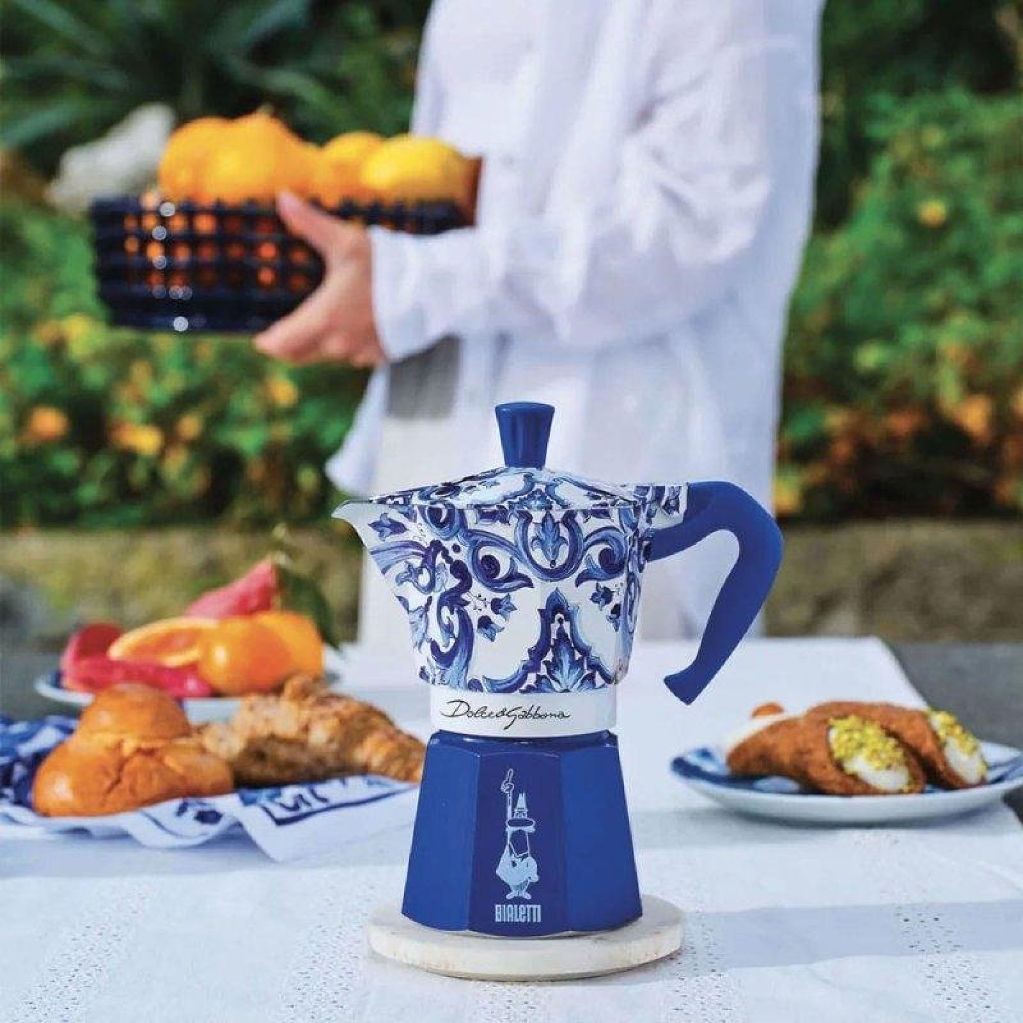 Una caffettiera Moka blu e bianca con motivo barocco su un tavolo con dolci e frutta.