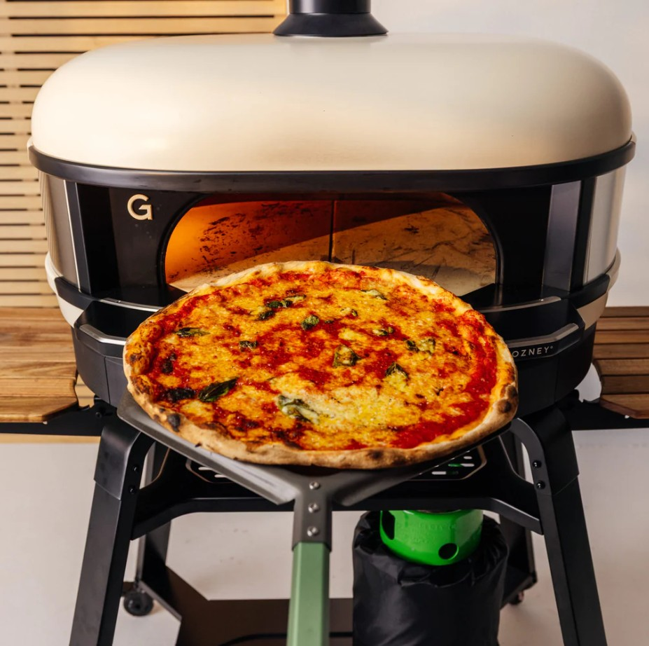 Eine frische Pizza auf einem Schieber vor einem modernen weißen Pizzaofen.