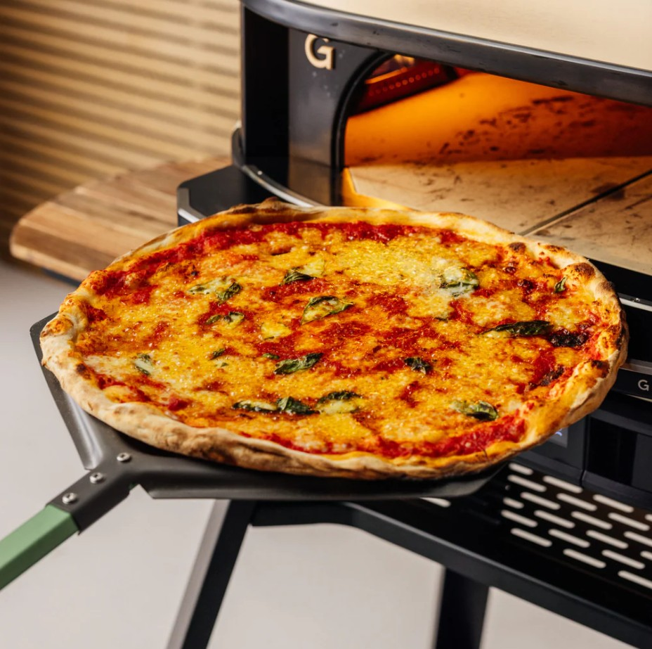 Eine frisch gebackene Pizza mit geschmolzenem Käse und Basilikum liegt auf einem Schieber vor einem Ofen.
