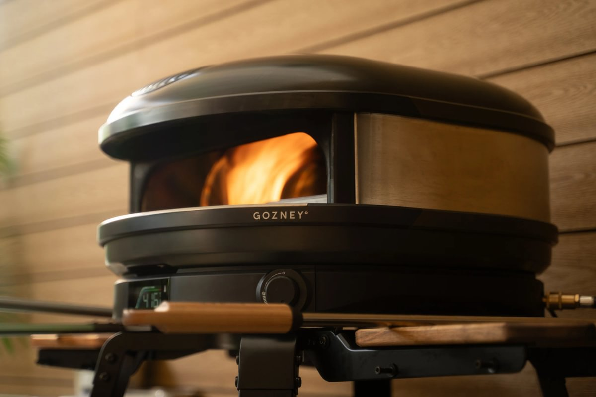 Ein schwarzer Gozney-Pizzaofen mit sichtbaren Flammen im Inneren steht vor einer Holzwand.