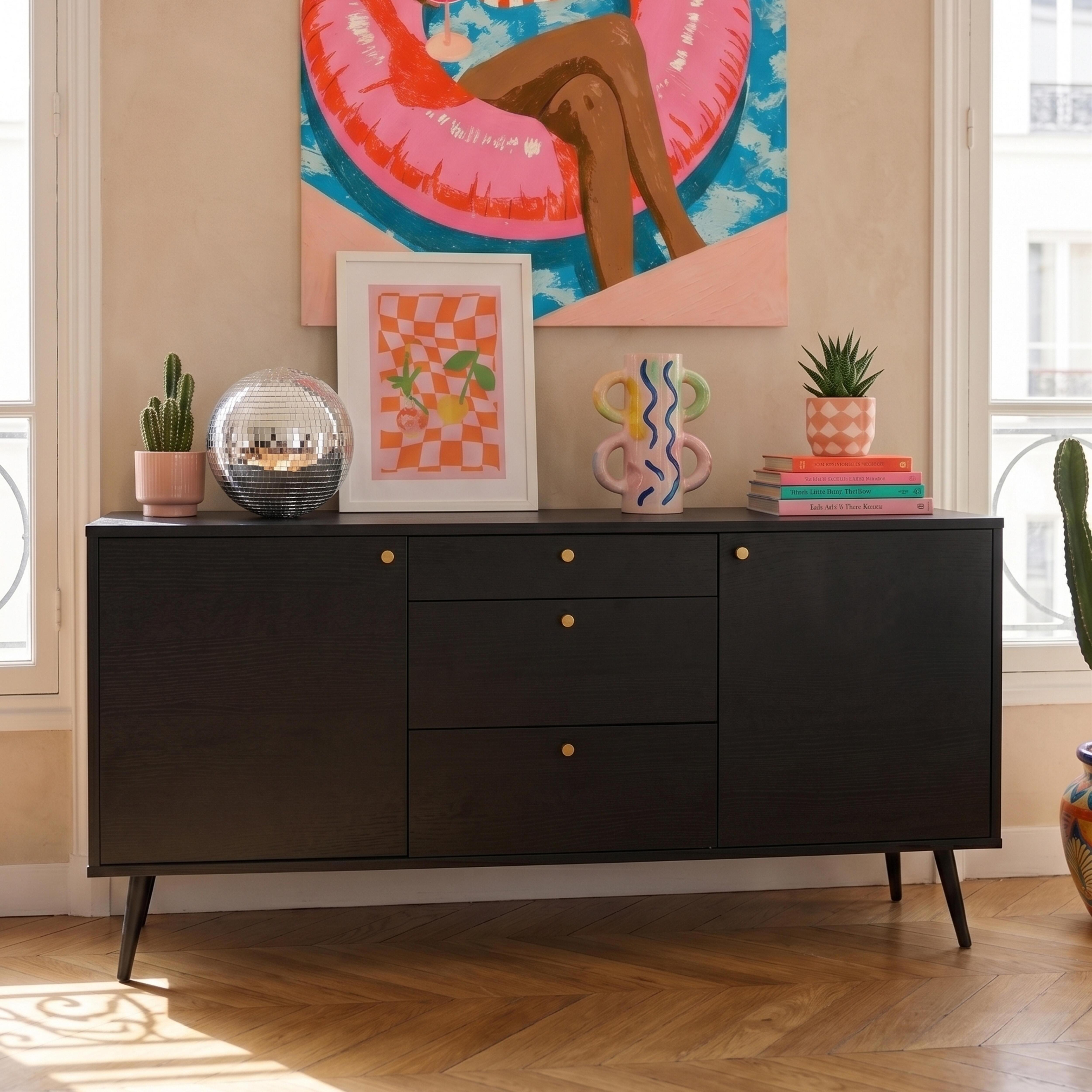 Ein schwarzes Sideboard mit Kunstwerken, einer Diskokugel und Kakteen davor, flankiert von Fenstern.