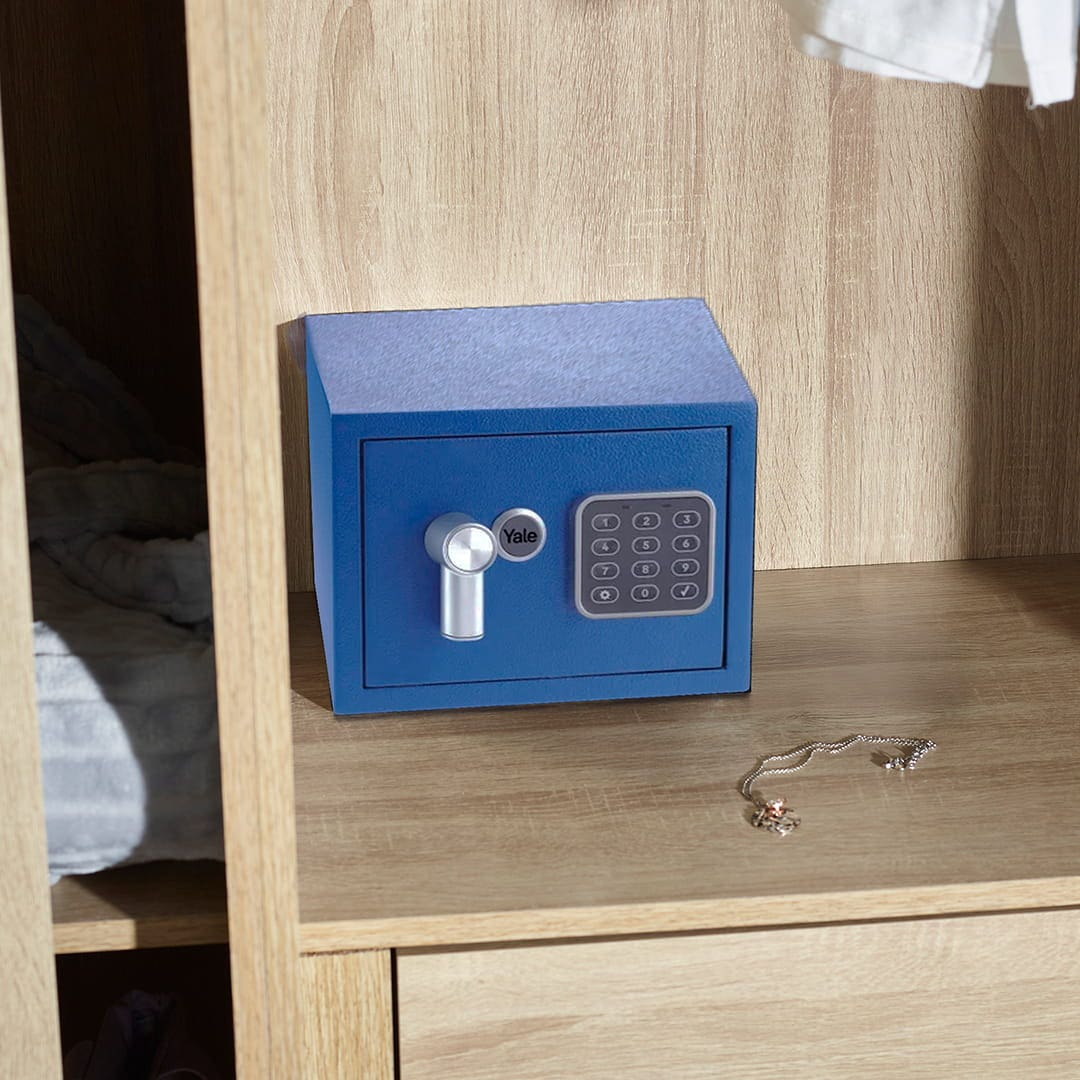 Una caja fuerte Yale azul con teclado y manija giratoria descansa en un estante de madera, con un collar cerca.