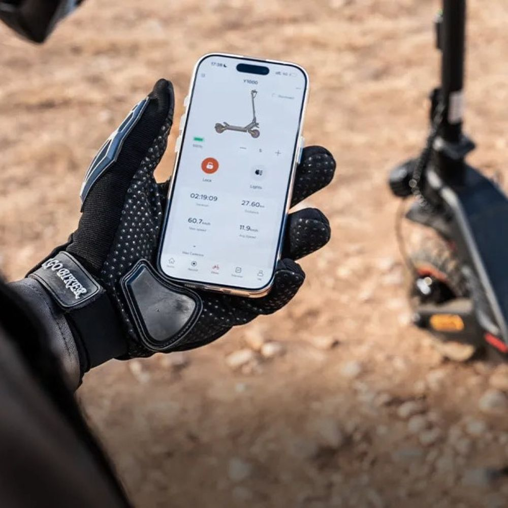 Una mano enguantada sostiene un smartphone con una aplicación de patinete eléctrico; al fondo, un patinete.