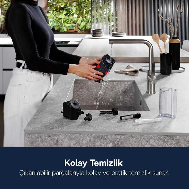 Bir kişi mutfak evyesinde, musluk suyu altında çıkarılabilir cihaz parçalarını yıkıyor.