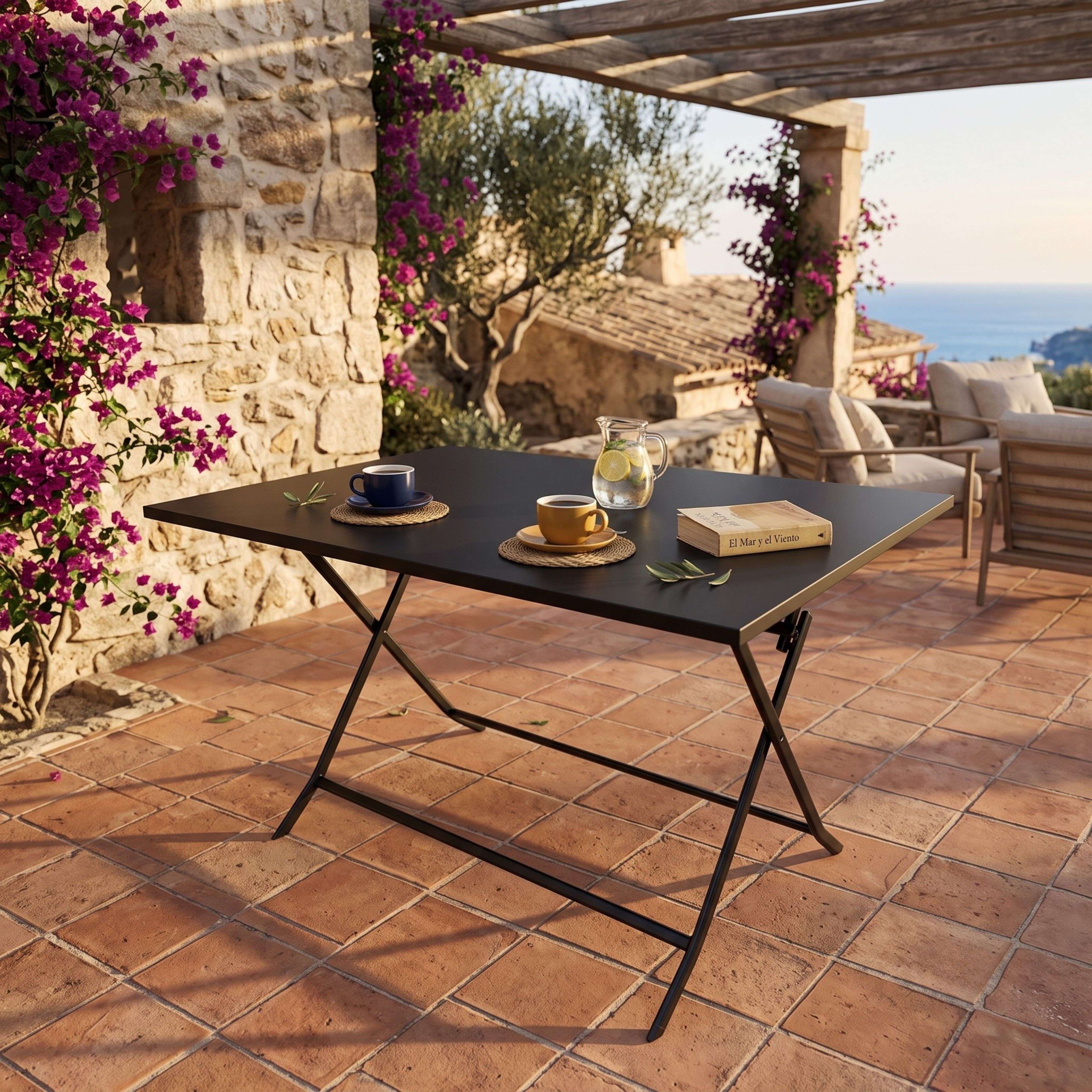 Mesa plegable negra en terraza con tazas de café, jarra de agua y libro, con vistas al mar.