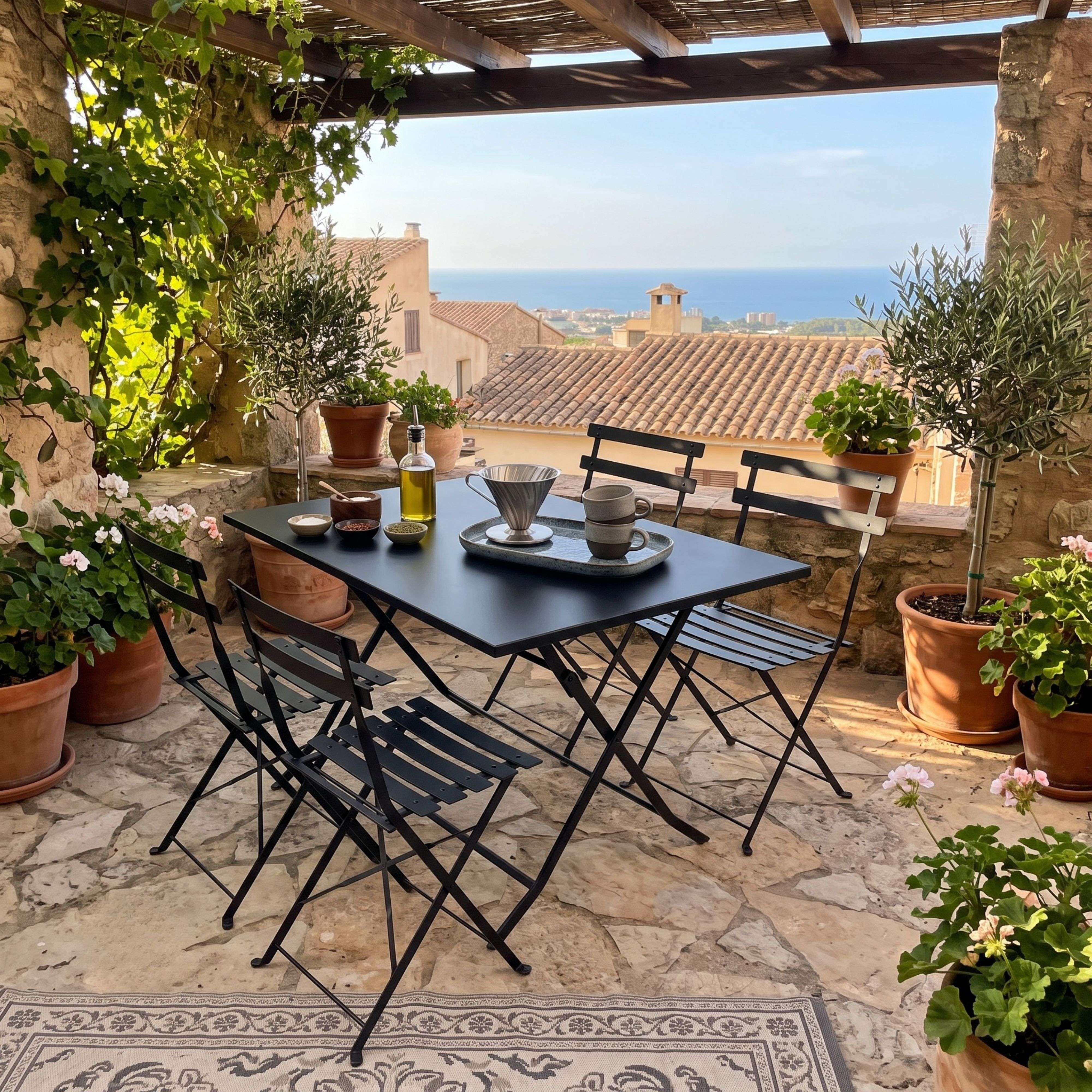 Una terraza de piedra con muebles de metal negro, olivos en macetas y vistas al mar.
