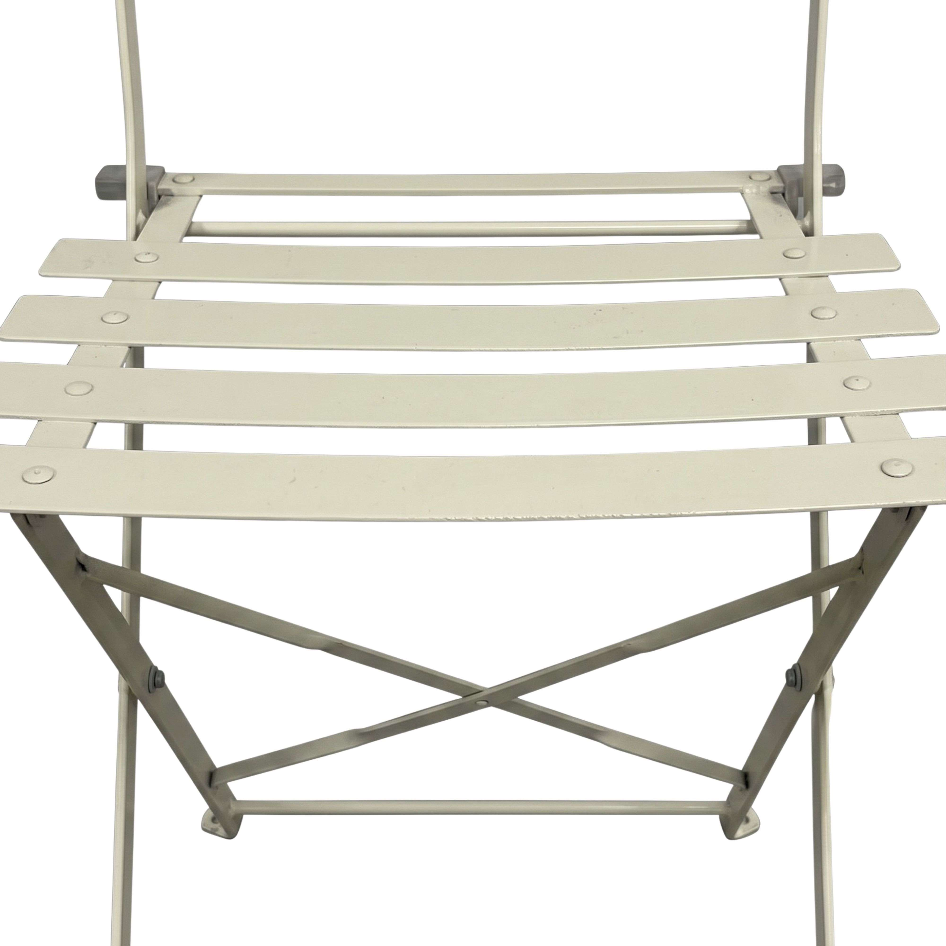 Primer plano del asiento y las patas cruzadas de una silla plegable de metal beige sobre fondo claro.
