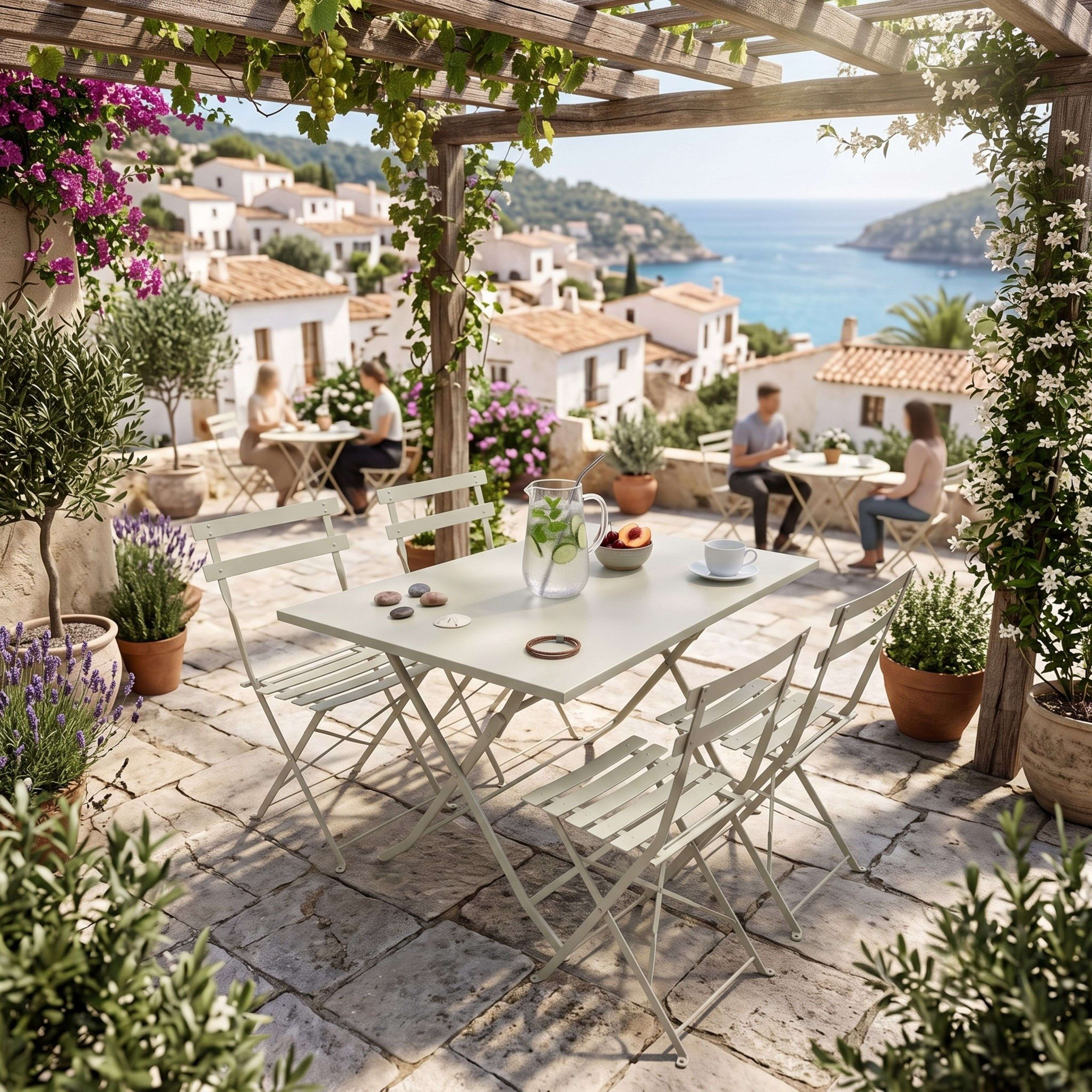 Un patio soleado con vistas al mar, mesas blancas, sillas, plantas y casas costeras.