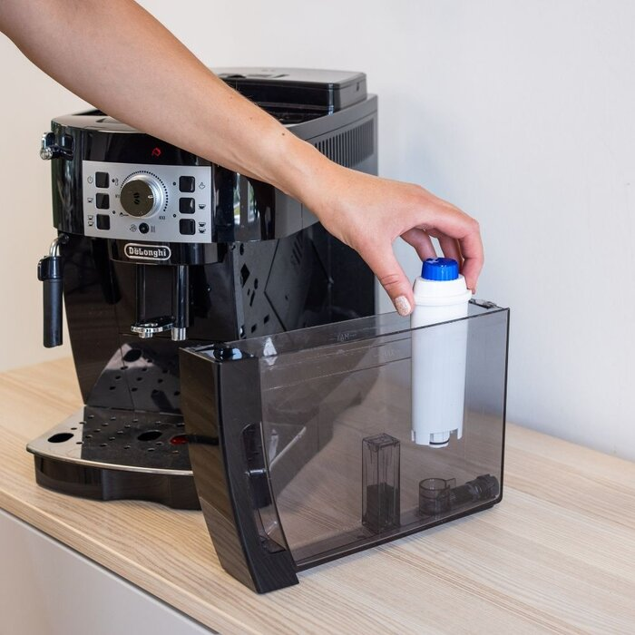 A hand places a water filter into the water tank of a black DeLonghi coffee machine.