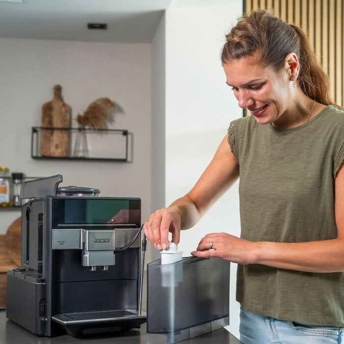 A woman inserts a water filter into the water tank of a black and silver espresso machine in a kitchen.