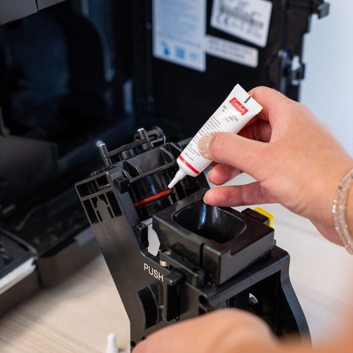 A hand applies grease to the red seal of a coffee machine brewing unit.