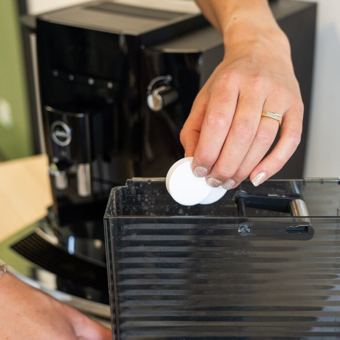 Een hand plaatst twee witte reinigingstabletten in het waterreservoir van een koffiezetapparaat.