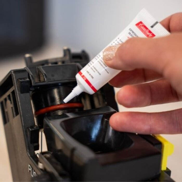 A hand applies silicone grease from a small tube onto the red gasket of a coffee machine.
